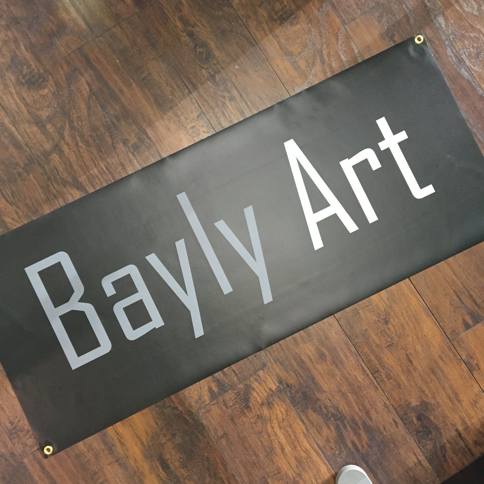 Bayly Art banner on a wood floor, black background with white lettering and grommets