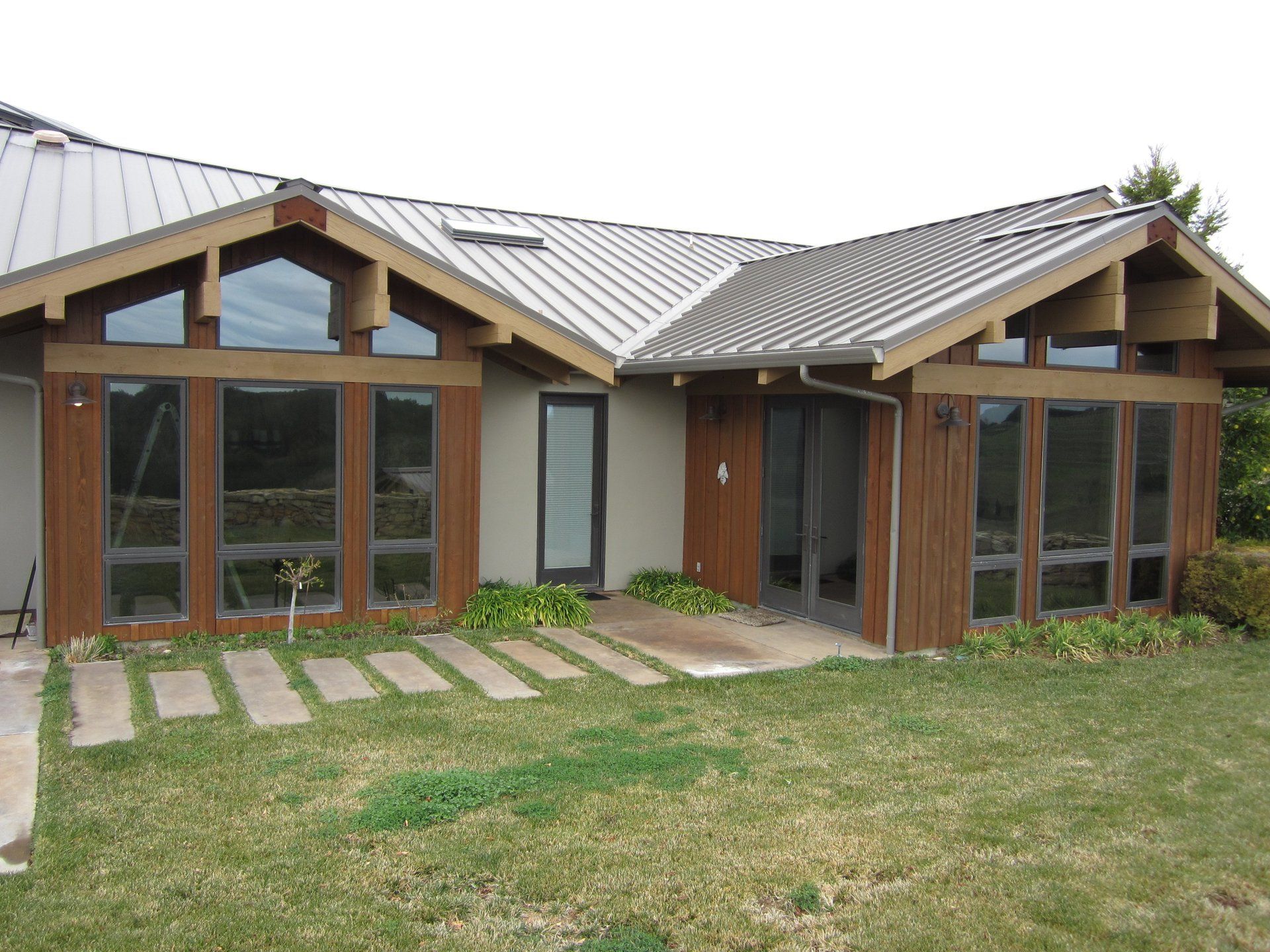 Modern house exterior with large windows, wood trim, and a metal roof, fronted by a grassy yard and stone path
