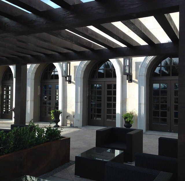 Shaded courtyard with arched windows, glass doors, and dark patio furniture under a wooden pergola