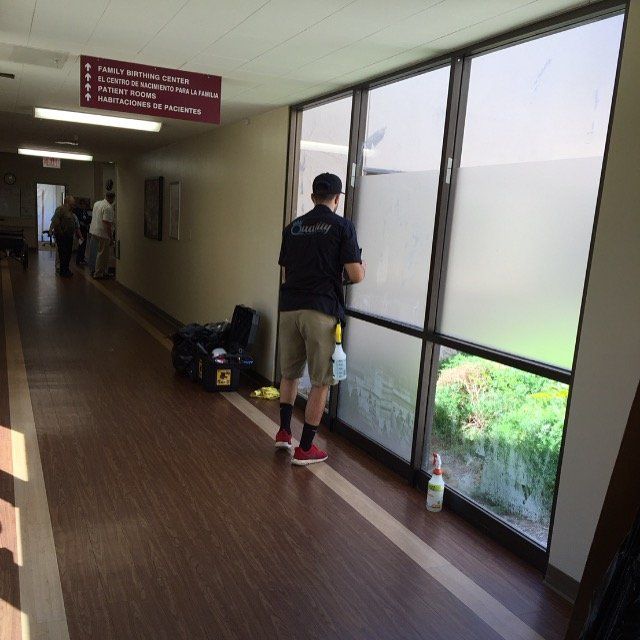 Person standing in a hallway by large windows, looking outside beside bags and a water bottle.