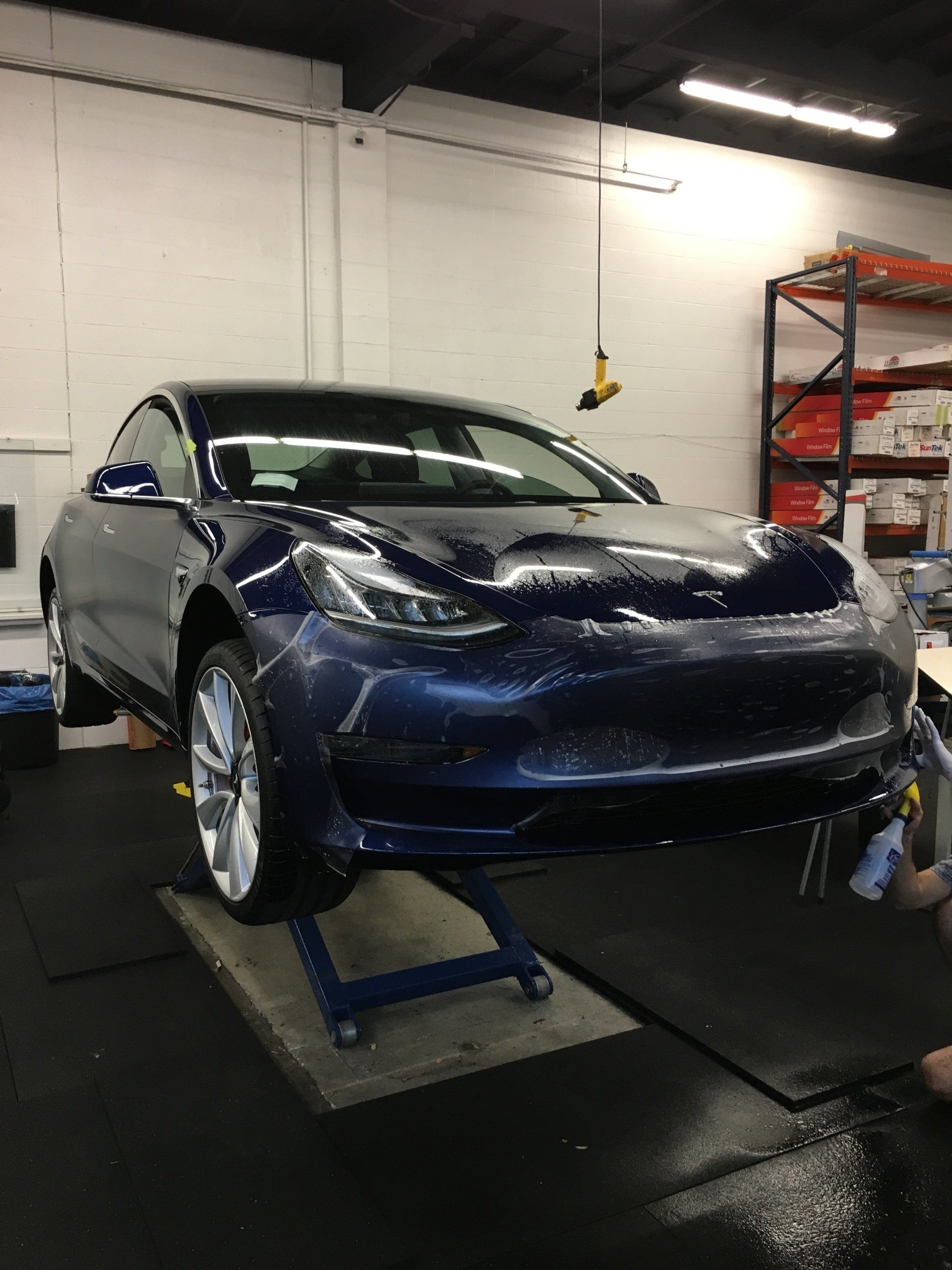 Blue car on a lift in an auto repair shop, with a rack of parts in the background.