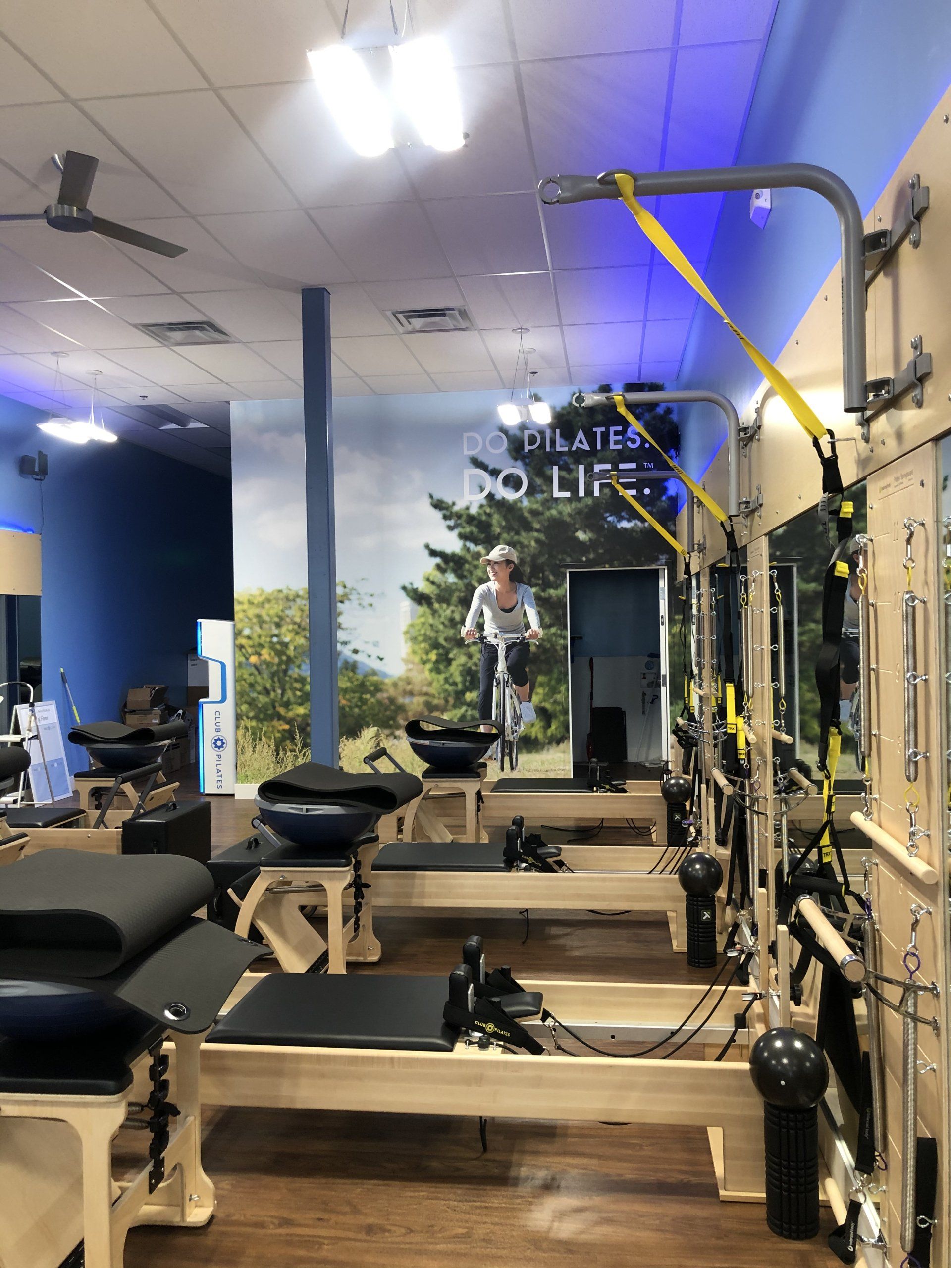 Pilates studio with reformer machines, suspension straps, mirrors, and overhead fans under blue lighting