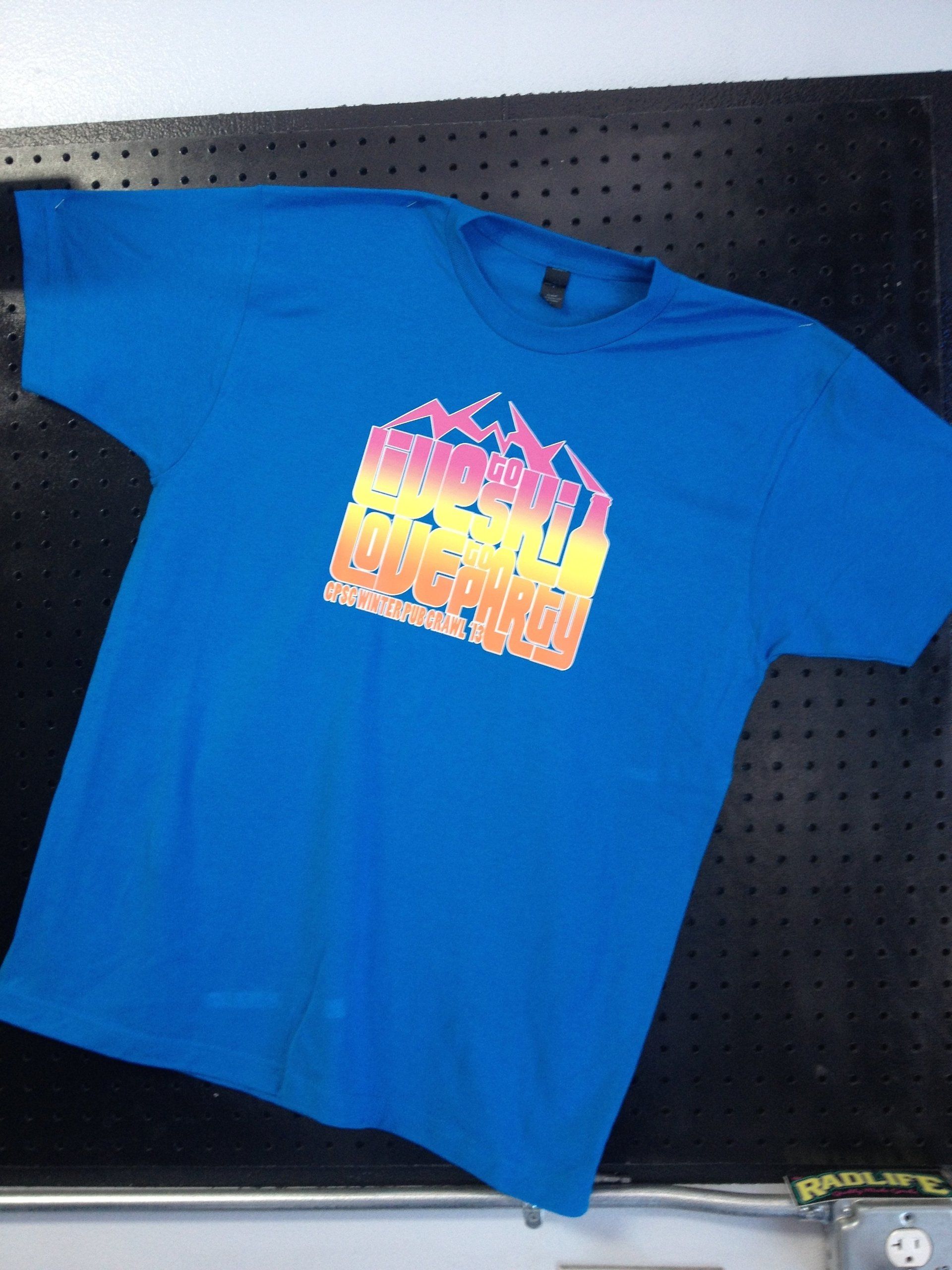 Blue T-shirt with a colorful graphic logo laid out on a dark pegboard display.