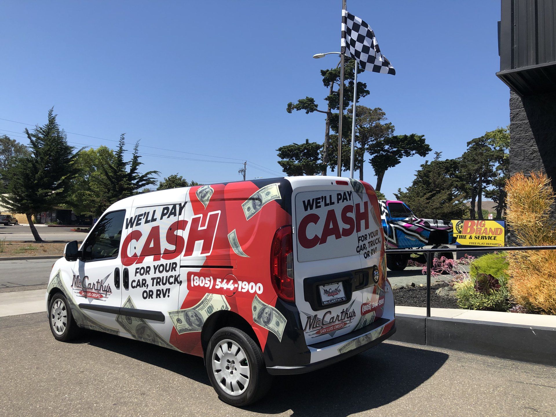 Wrapped van advertising cash for cars and welfare, parked by a building under a checkered flag.