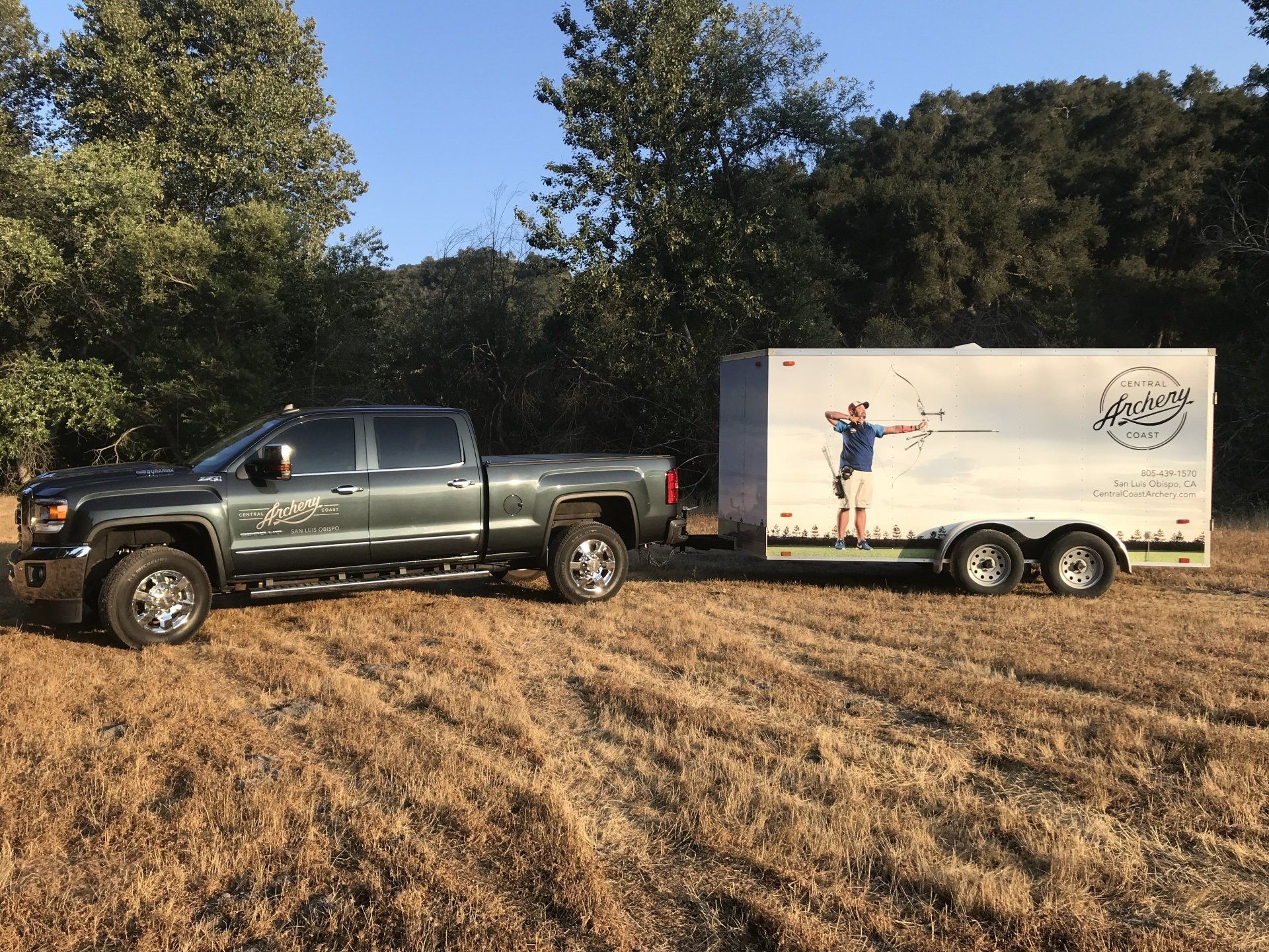 Green pickup truck towing a white trailer in a grassy field under trees and blue sky
