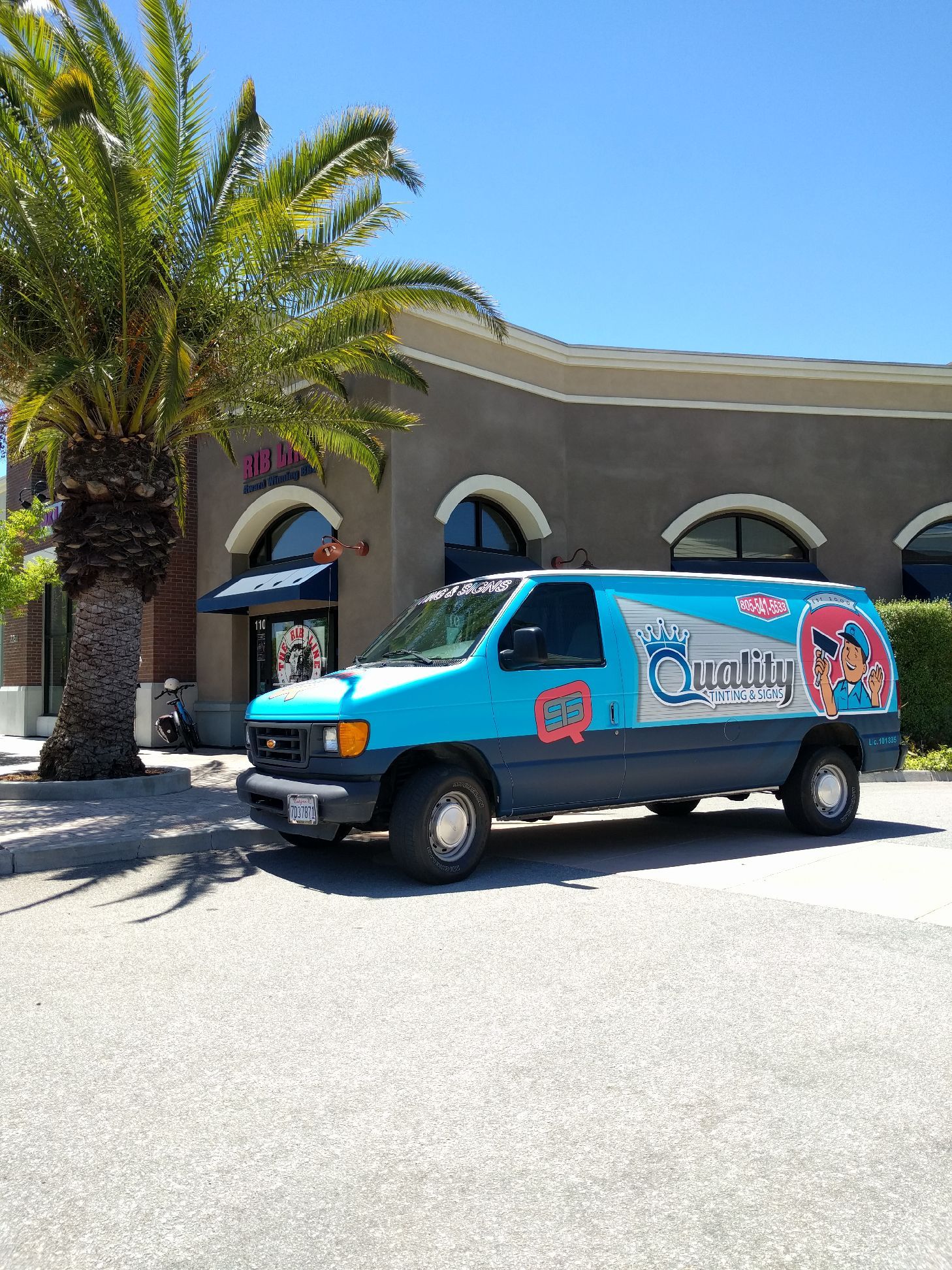 Turquoise van with logo parked beside a palm tree and building under a bright blue sky.