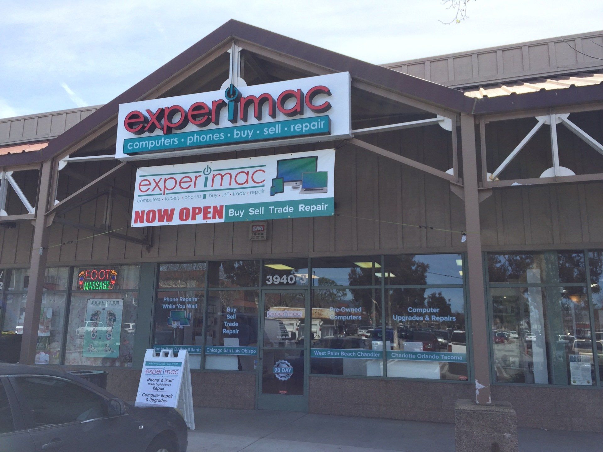 Exterior of an Experimac storefront with “Now Open” signage in a shopping center.