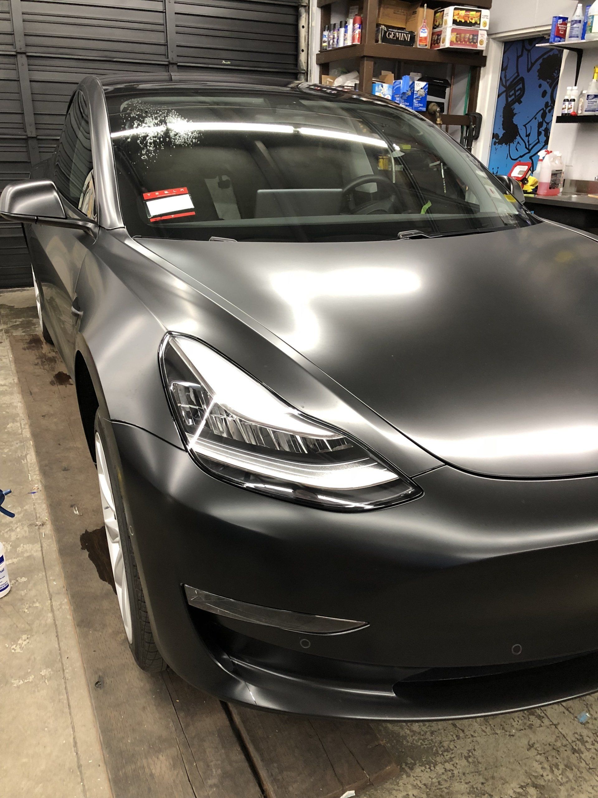 Matt black Tesla sedan parked in a garage, front-left view, with shelves and tools in the background