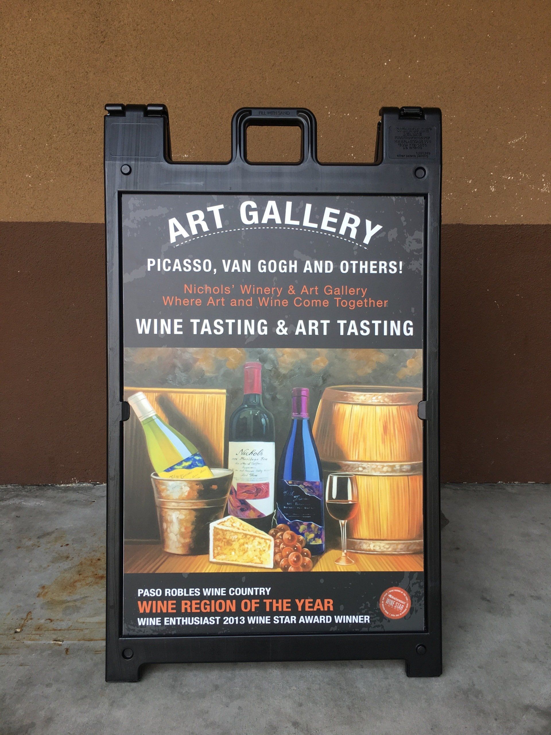 Black sandwich-board sign for an art gallery wine tasting with colorful painted wine bottles and barrels.