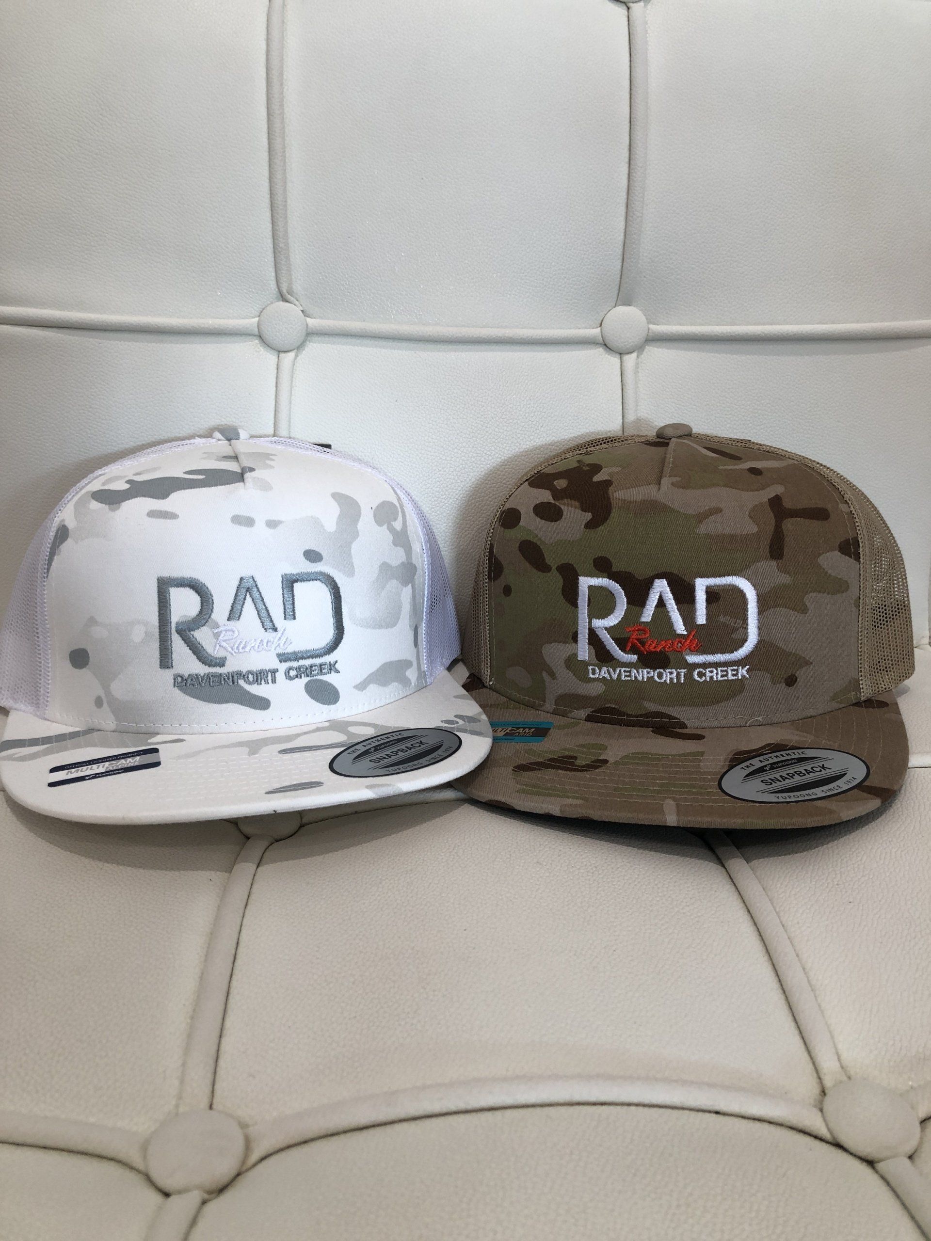 Two RAD logo baseball caps, one white and one brown, on a tufted white chair