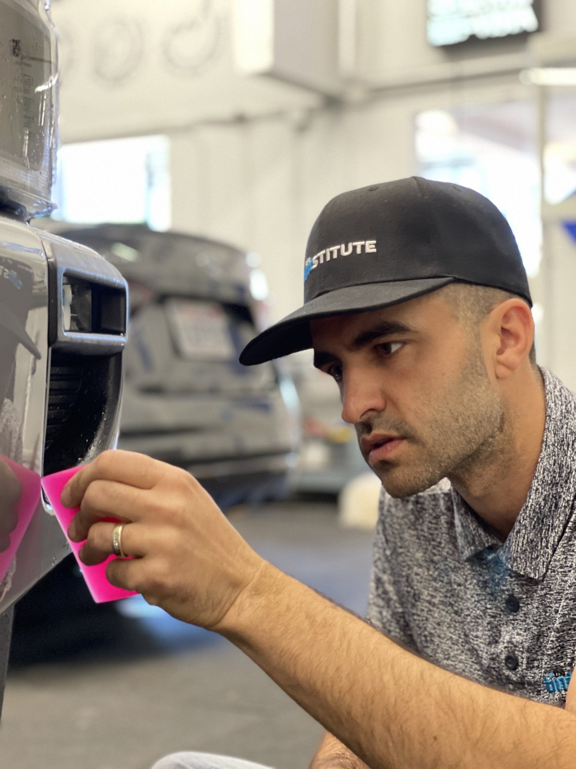Man in black cap carefully applying pink tape to a car part in a workshop