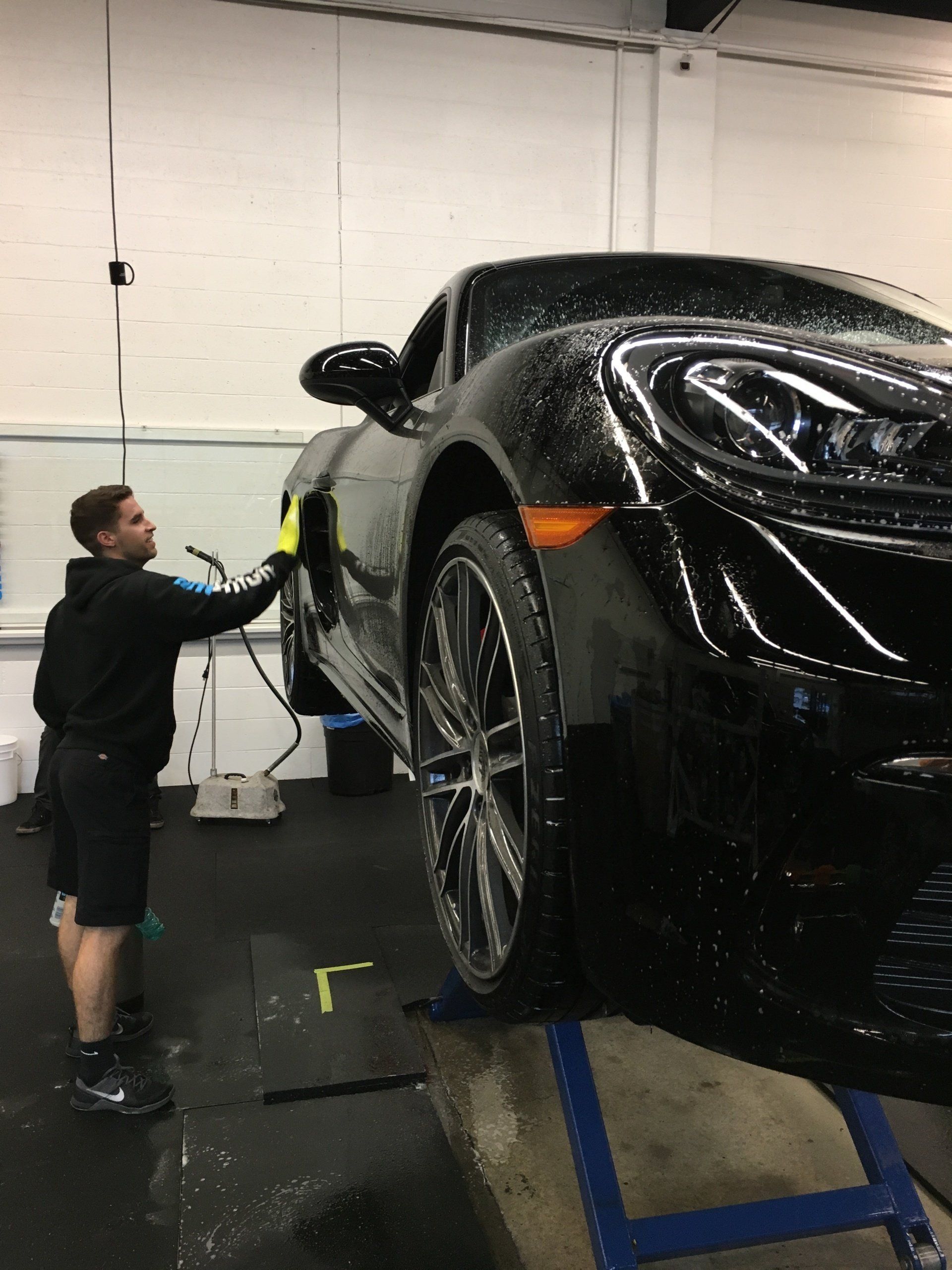 Person cleaning a black sports car in a garage, standing beside the front wheel on a lift.