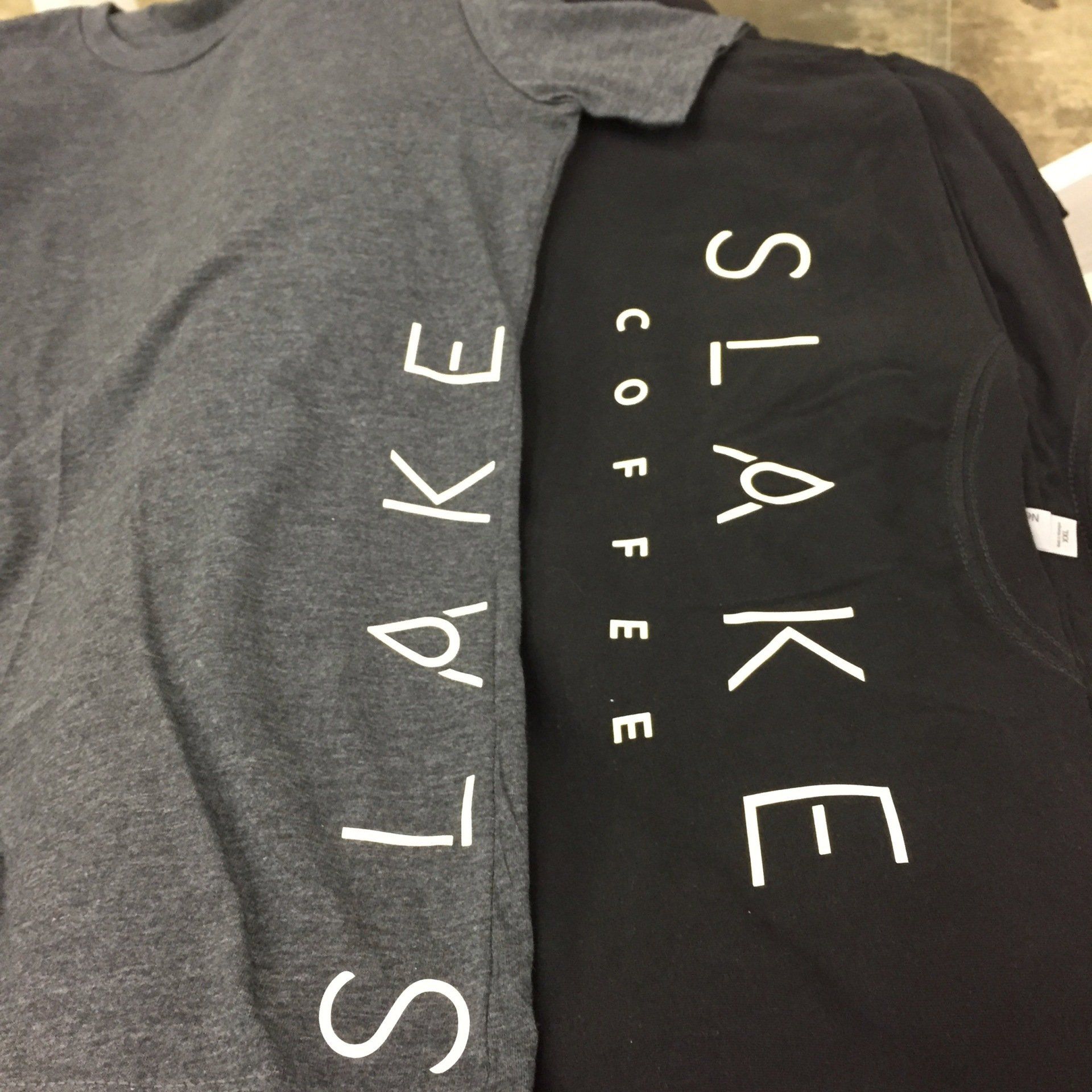 Two folded SLAKE coffee T-shirts, one gray and one black, with white lettering