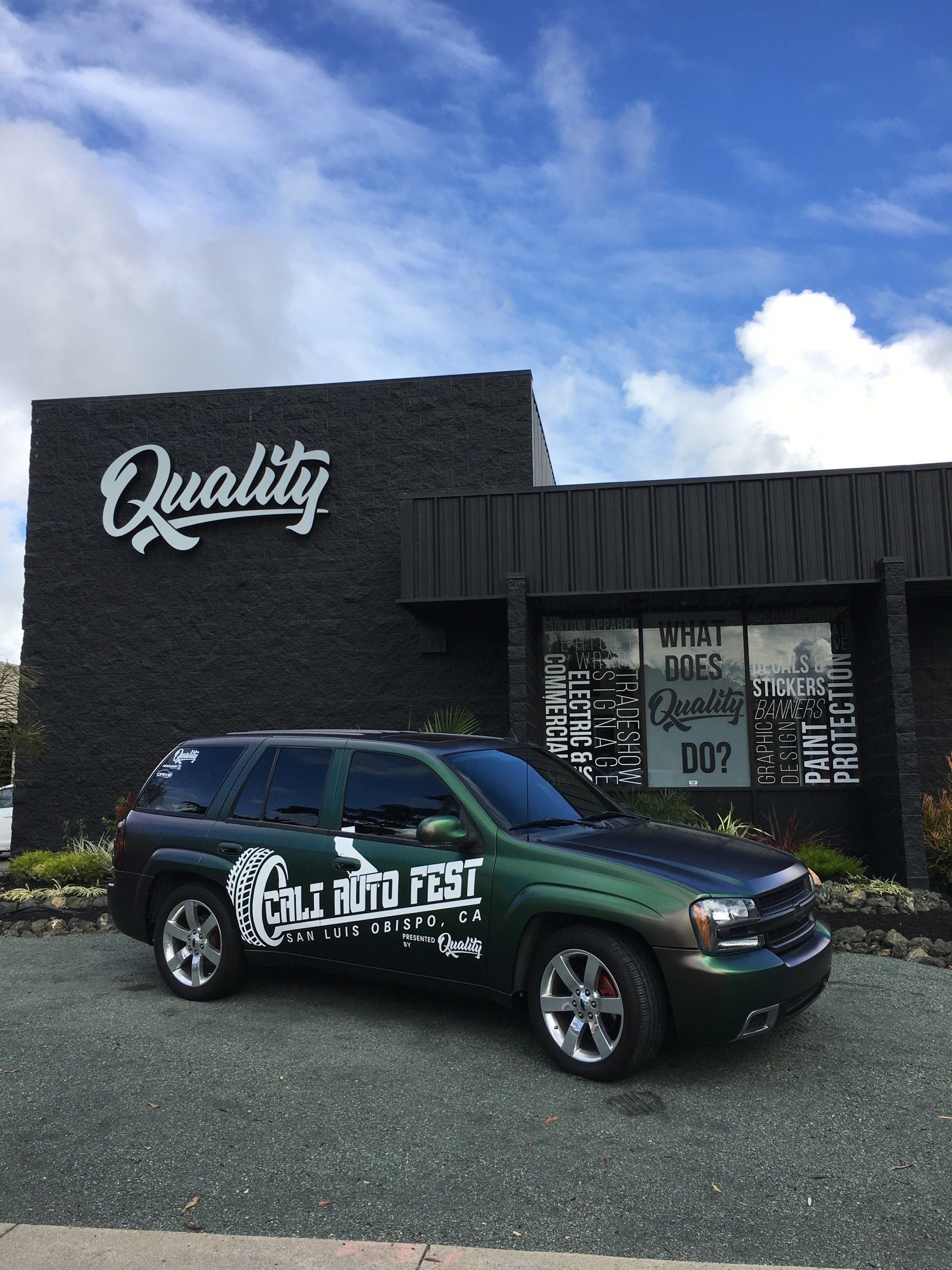 Custom pickup truck parked outside a black “Quality” building under a cloudy sky.