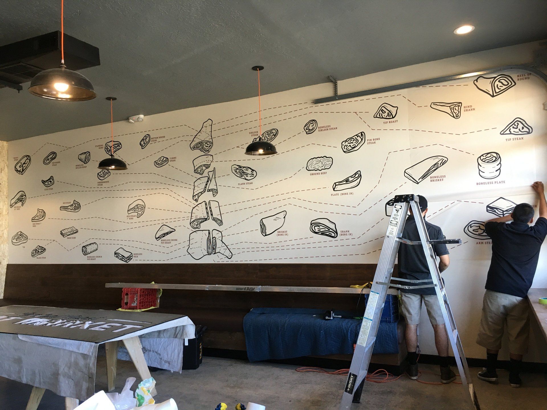 Person painting a mural on a wall in a restaurant with black-and-white doodles and a ladder nearby
