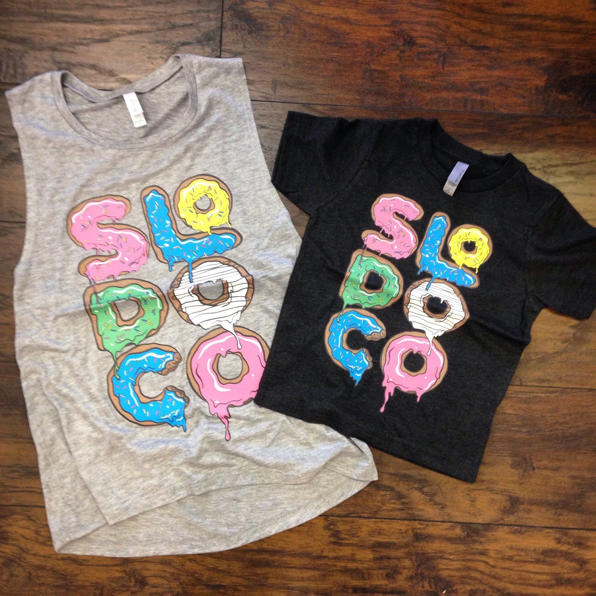 Two T-shirts on a wooden surface, one gray and one black, with colorful donut letters spelling “SLOP CO.”