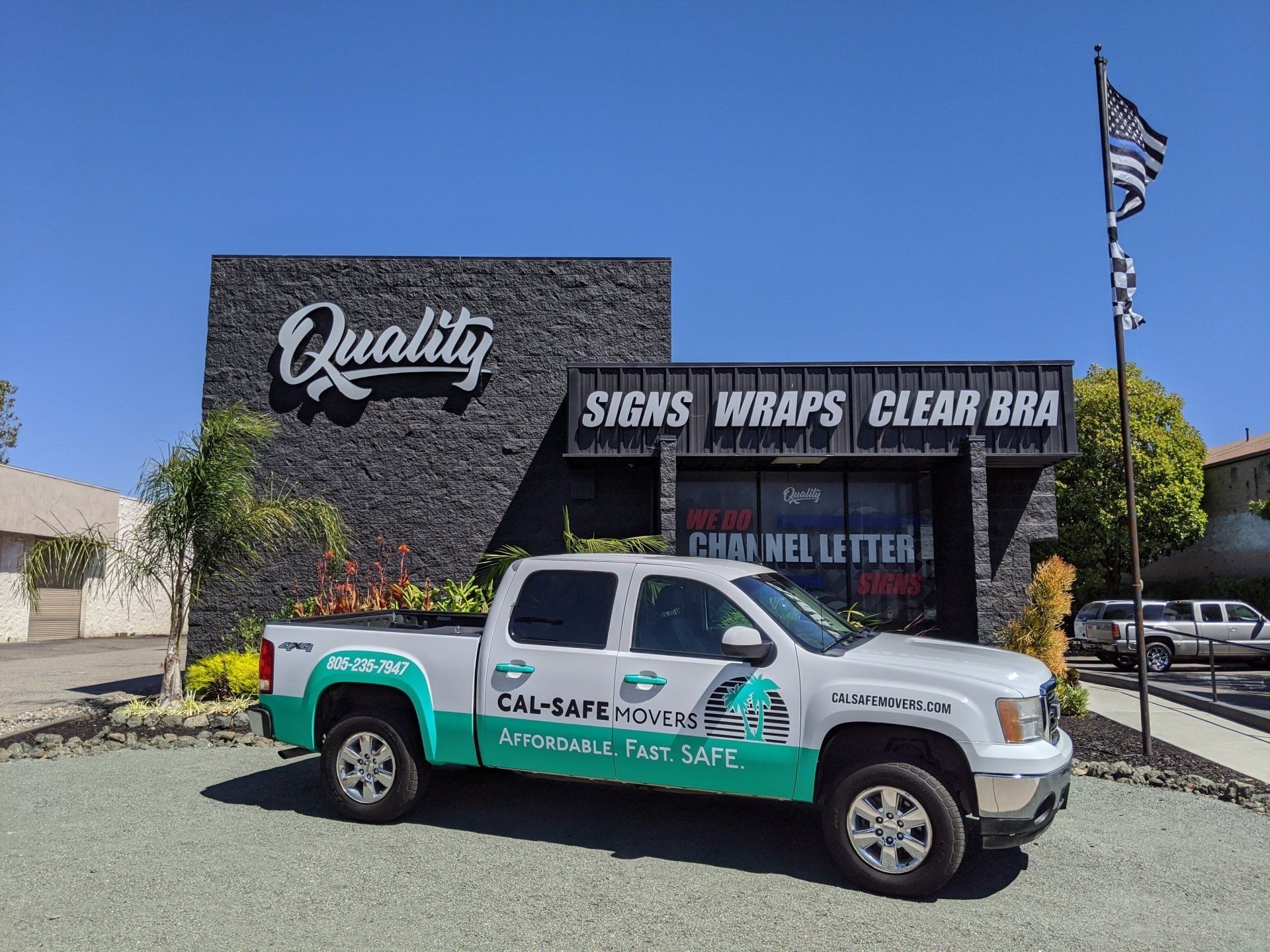 White pickup with turquoise graphics parked outside a “Quality” storefront under a clear blue sky.