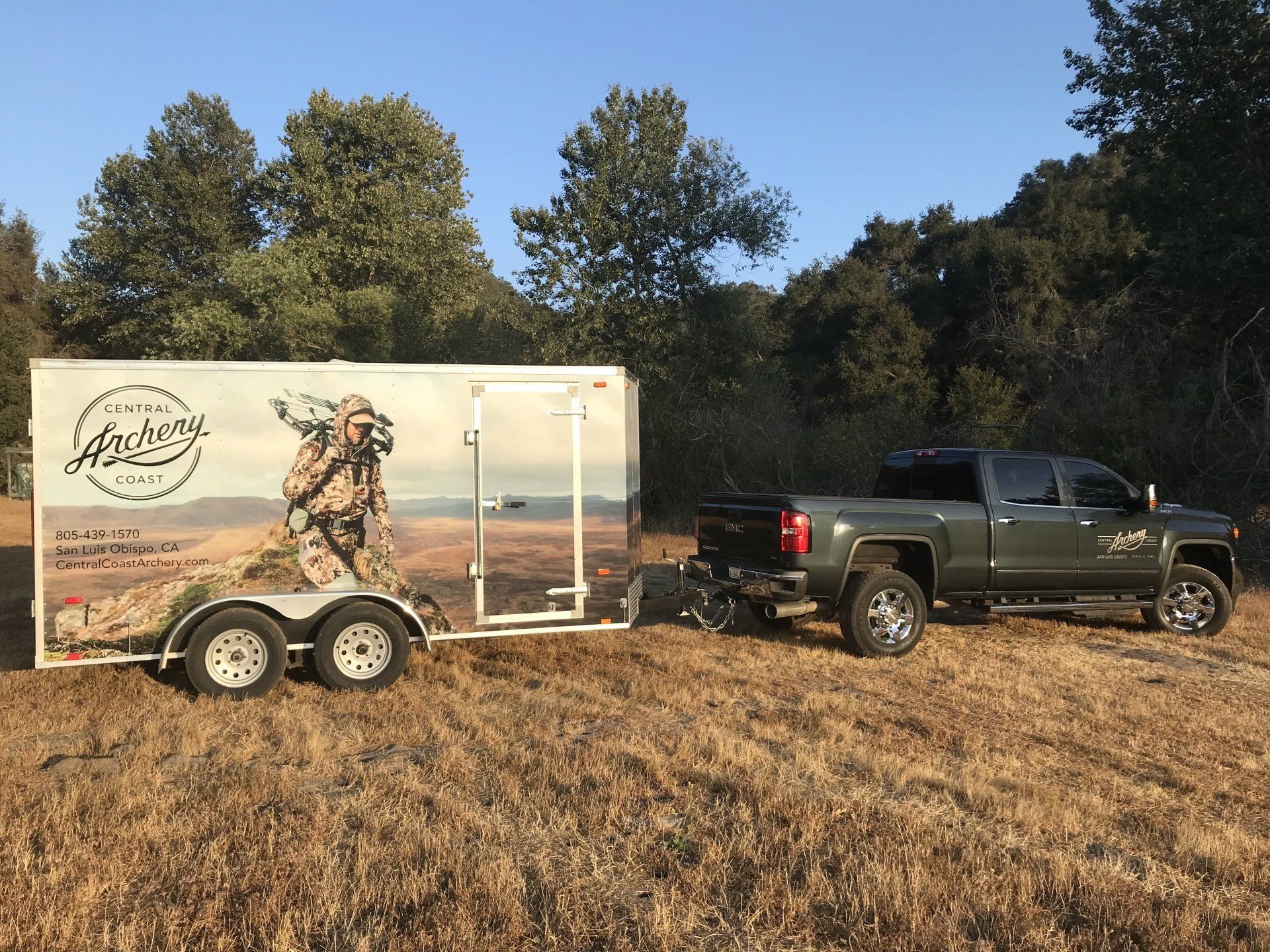 Dark pickup truck towing a hunting-themed trailer across a grassy field at sunset