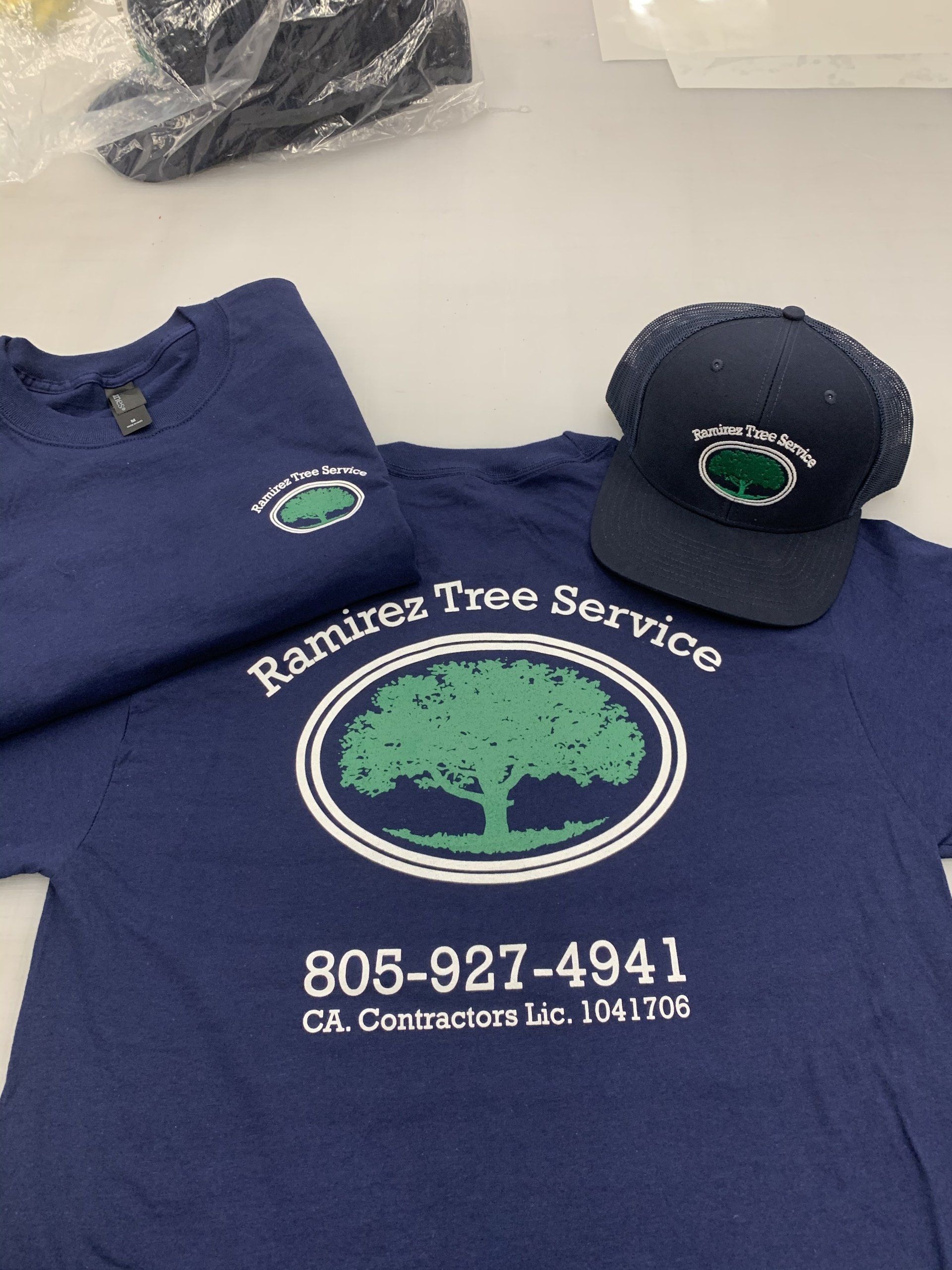 Blue Ramiroz Tree Service shirt and cap with tree logo and phone number on a white table