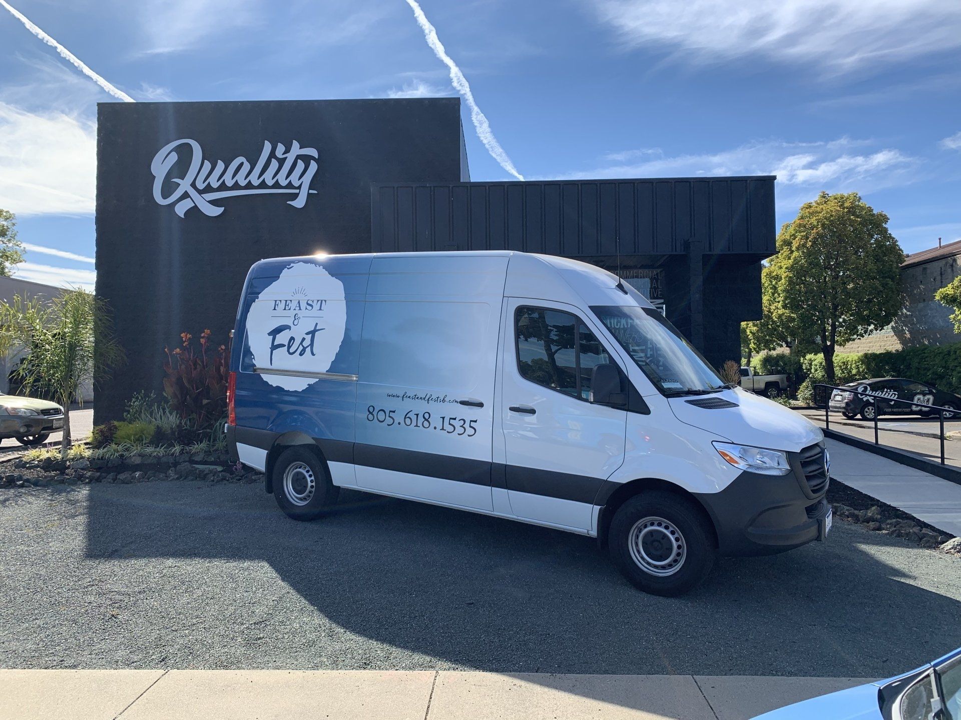 White delivery van parked outside a building with a “Quality” sign under a blue sky.