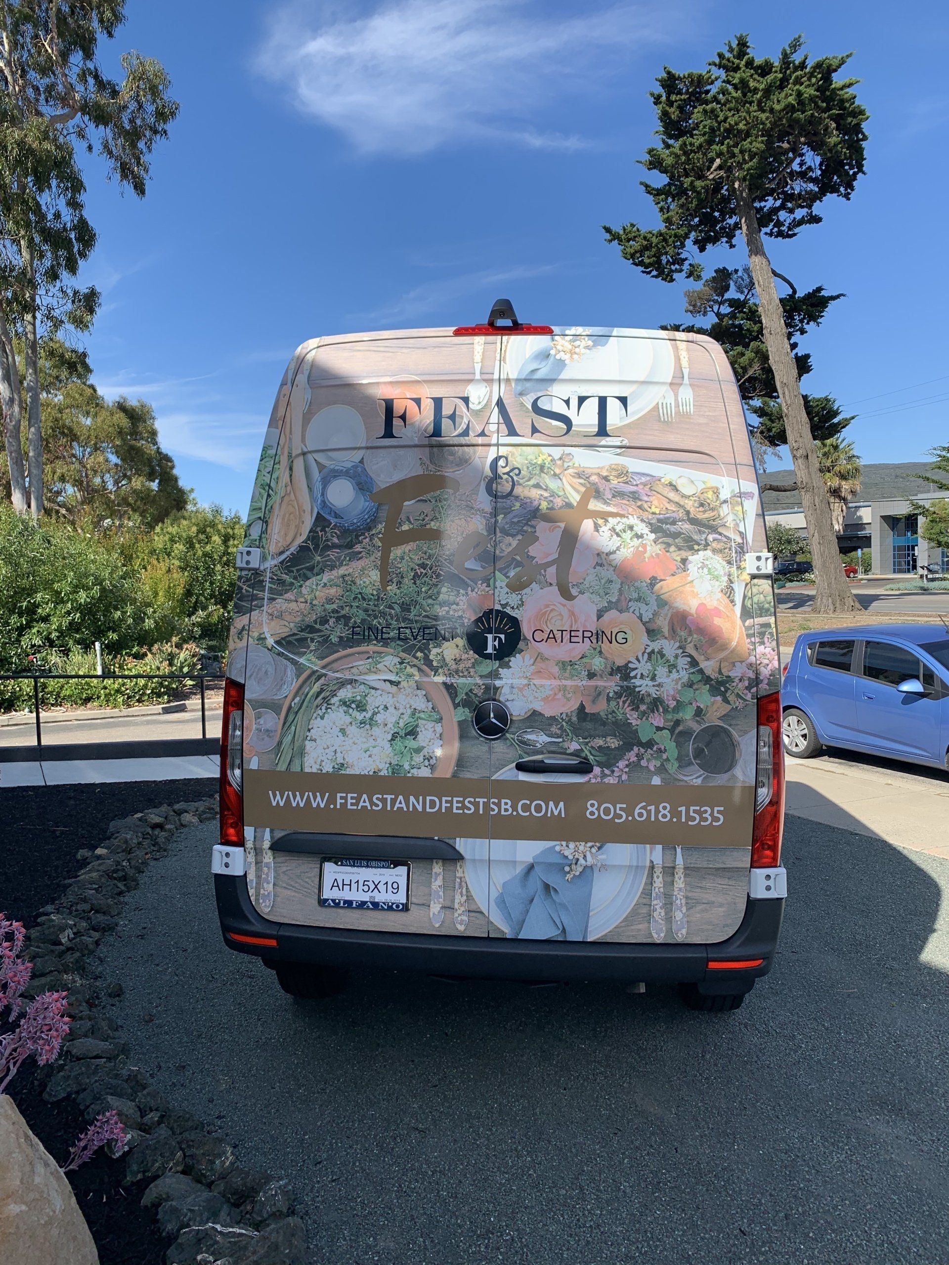 Decorated van parked outdoors under blue sky, with colorful map-like artwork on the rear door.