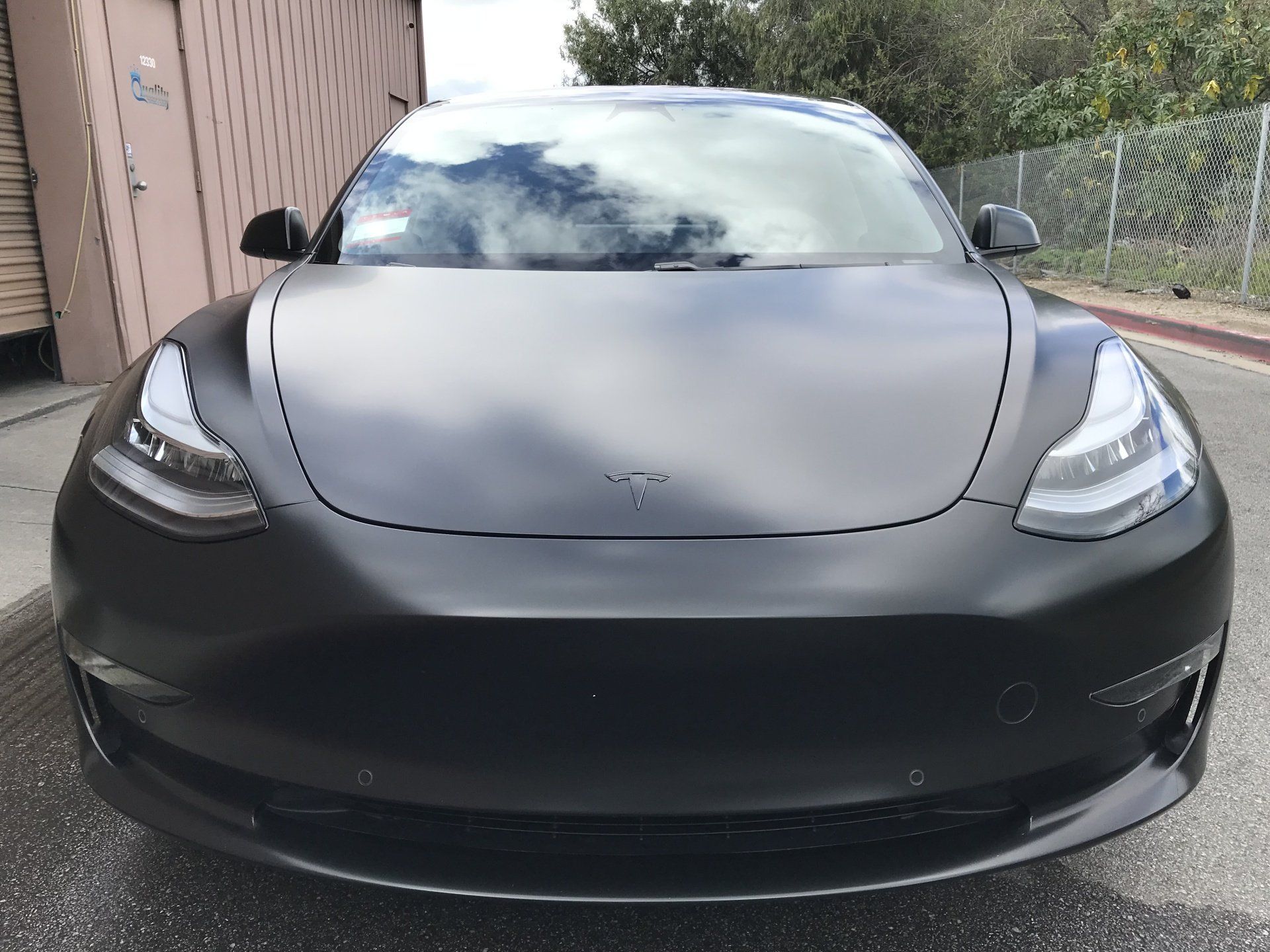 Front view of a black Tesla parked outdoors beside a house and fence