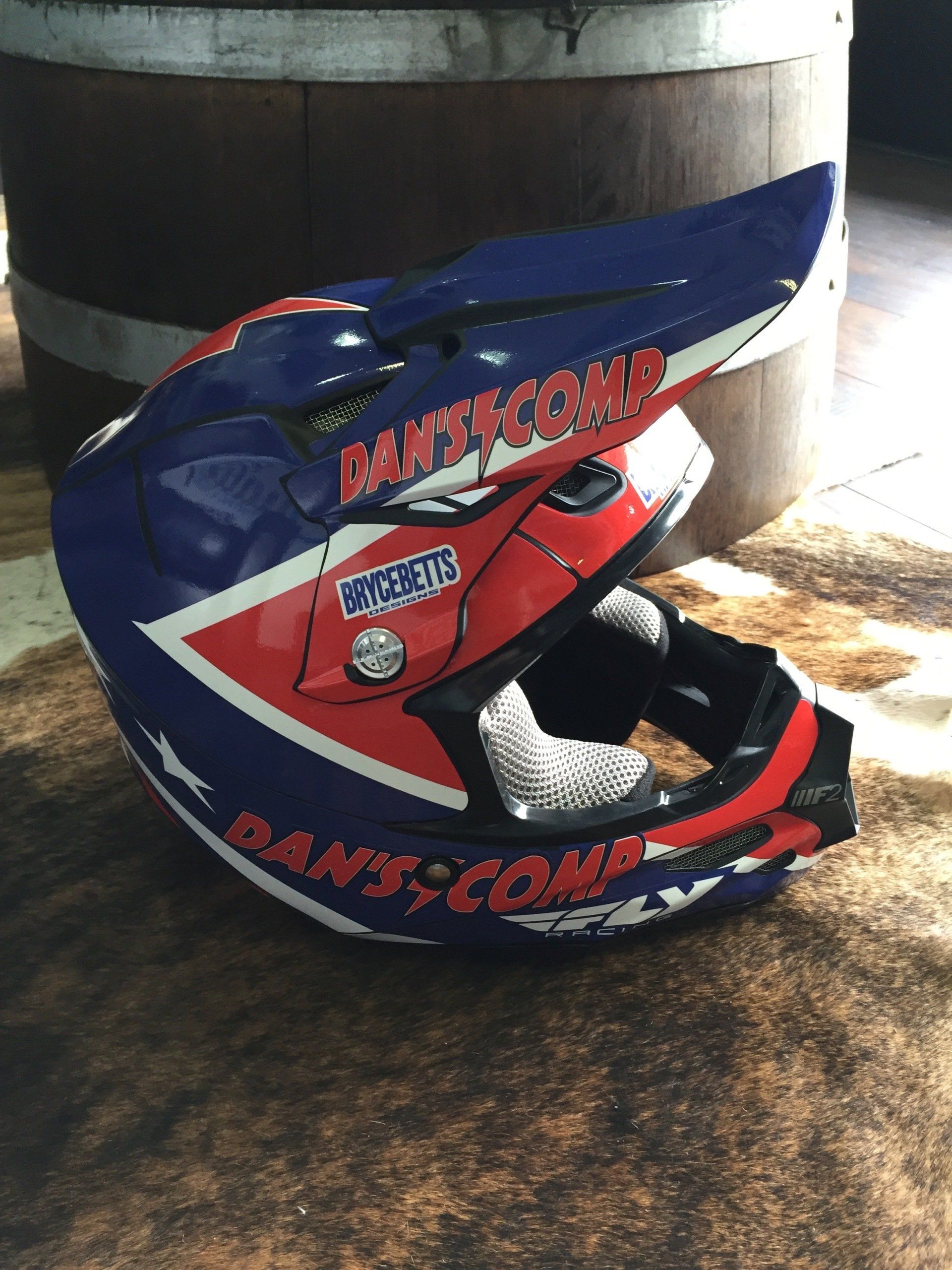 Blue, red, and white motocross helmet with sponsor logos resting on a floor near a large barrel.