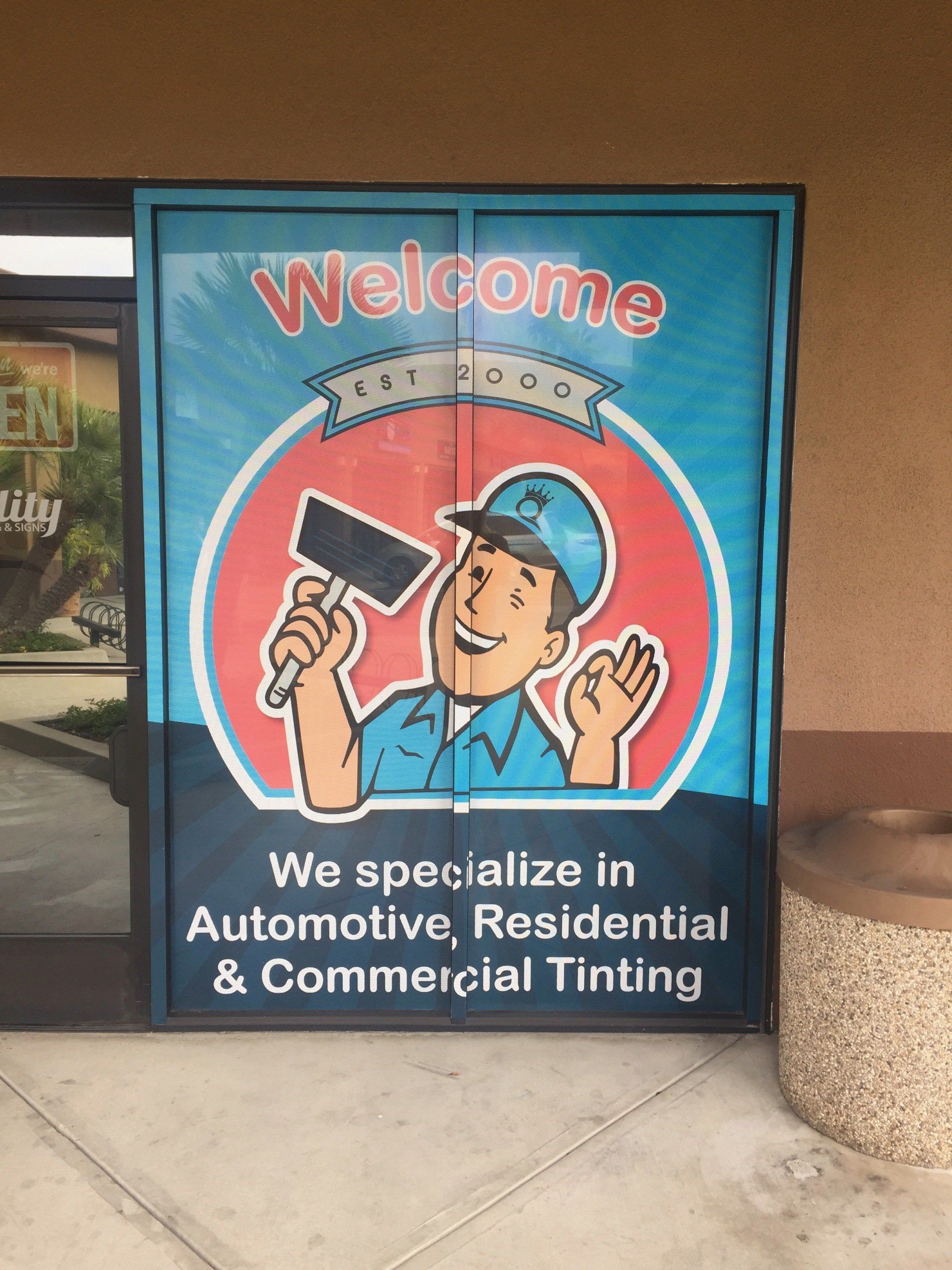 Blue storefront sign reading “Welcome” for automotive, residential and commercial tinting, with a cartoon worker painting.