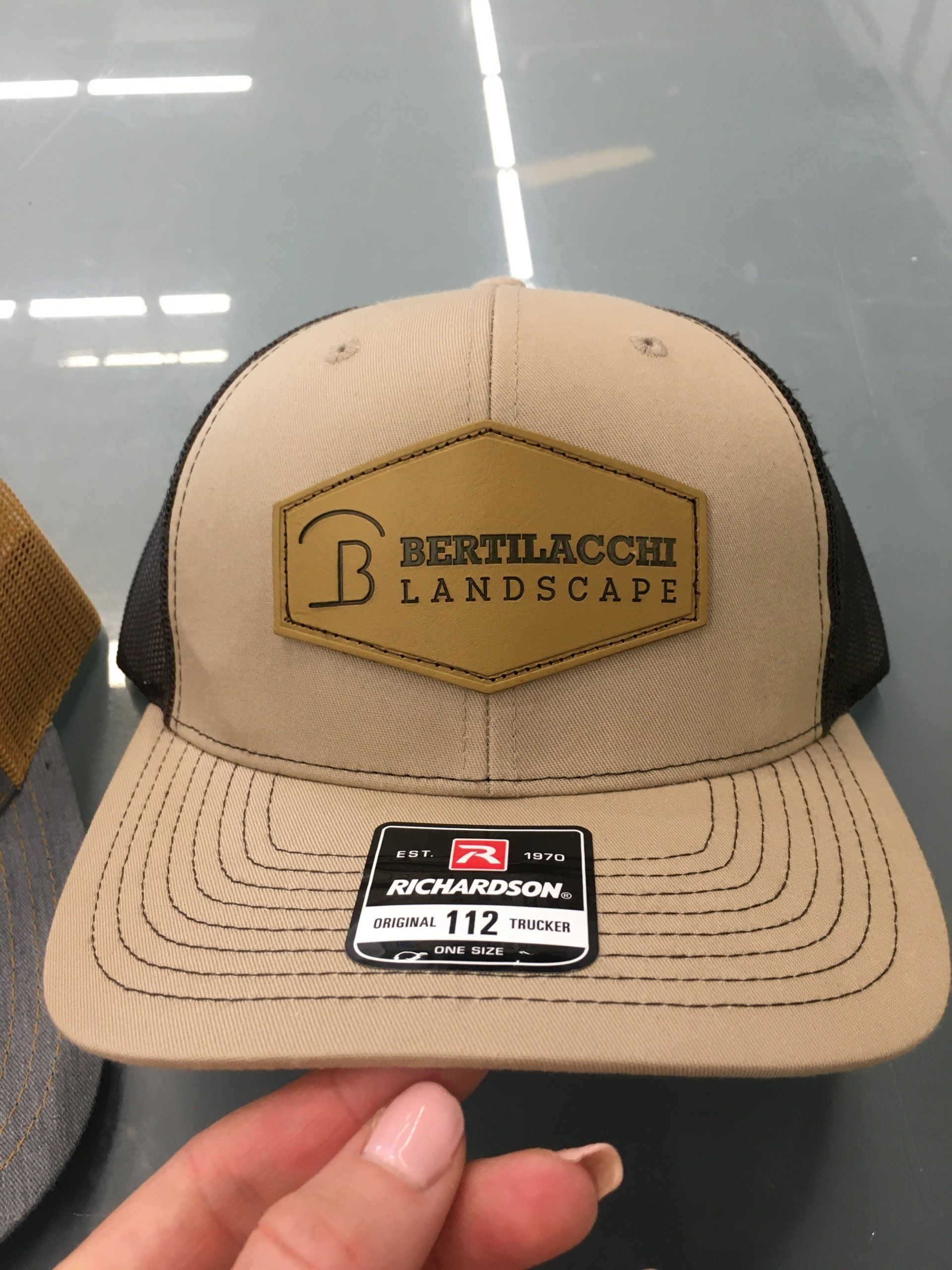 Tan mesh-and-black trucker hat with a “Bethlacco Landscape” patch, held by a hand.