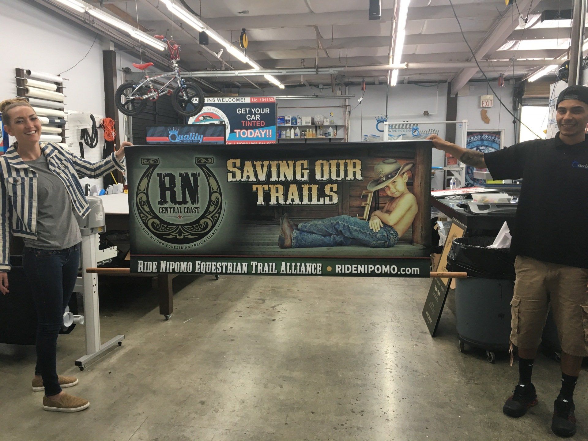 Two people hold a large “Saving Our Trails” banner in a workshop or garage.