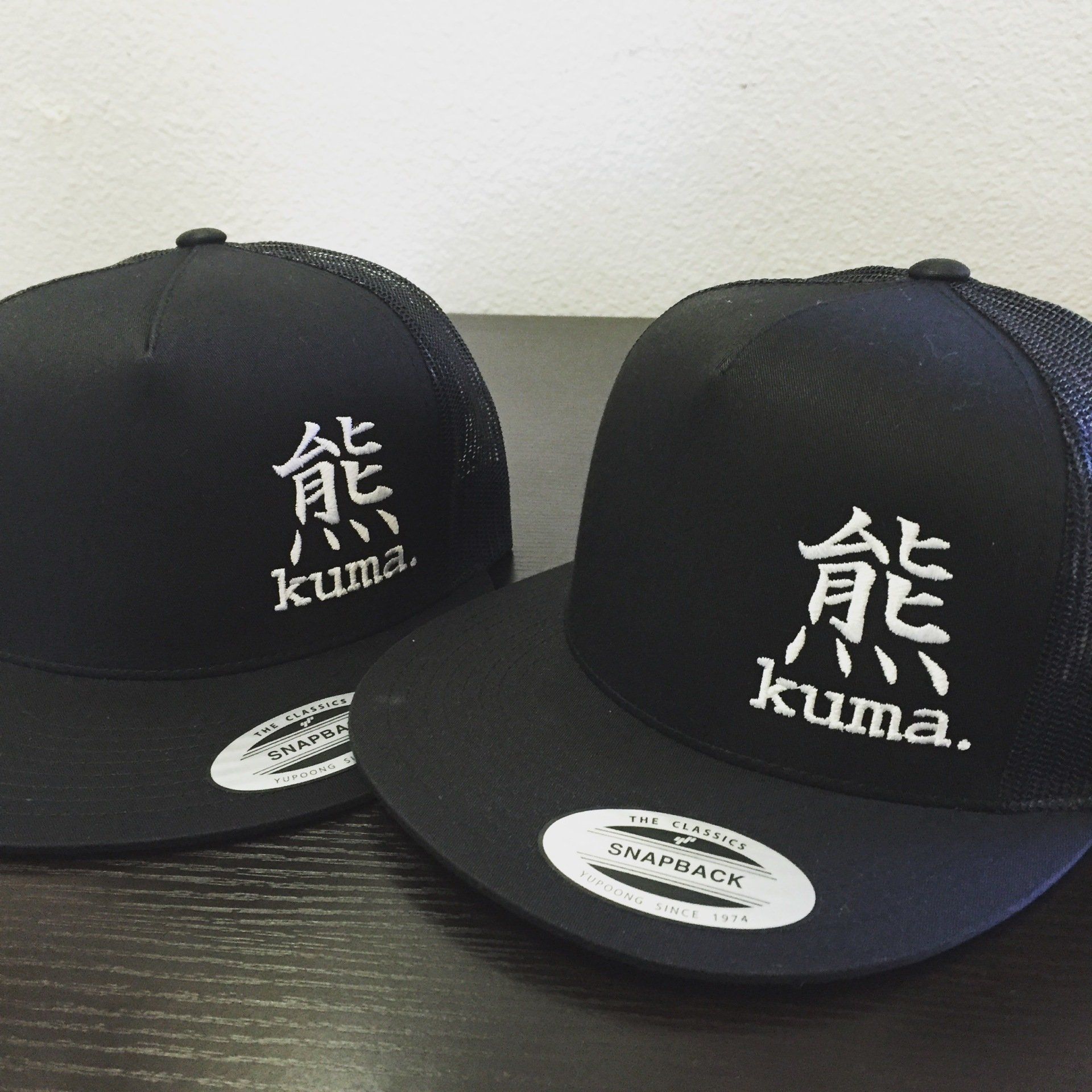 Two black Kuma baseball caps with white logo lettering, displayed side by side on a flat surface.