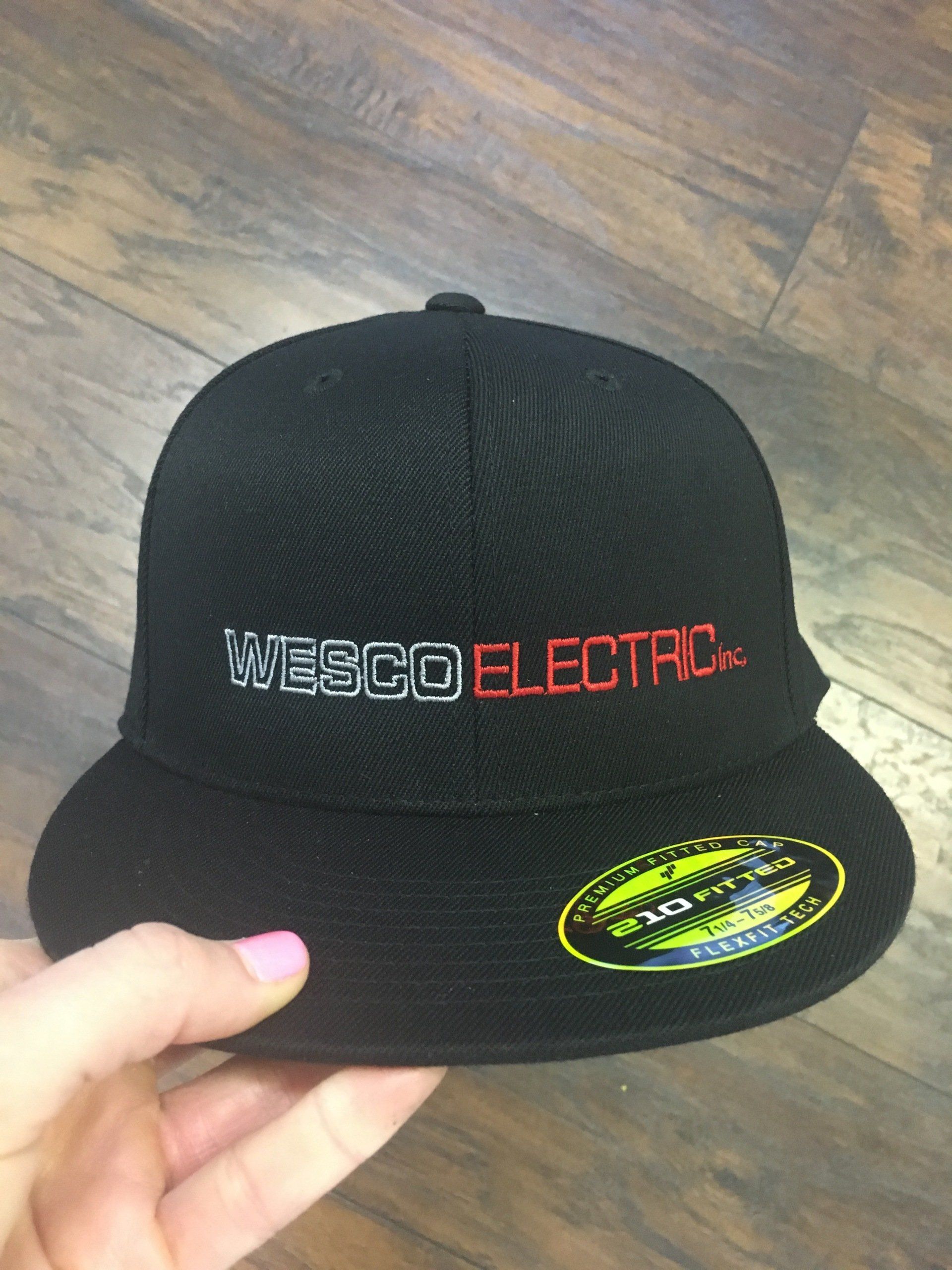 Black Wesco Electric cap with embroidered logo and neon oval sticker, held over a wooden floor