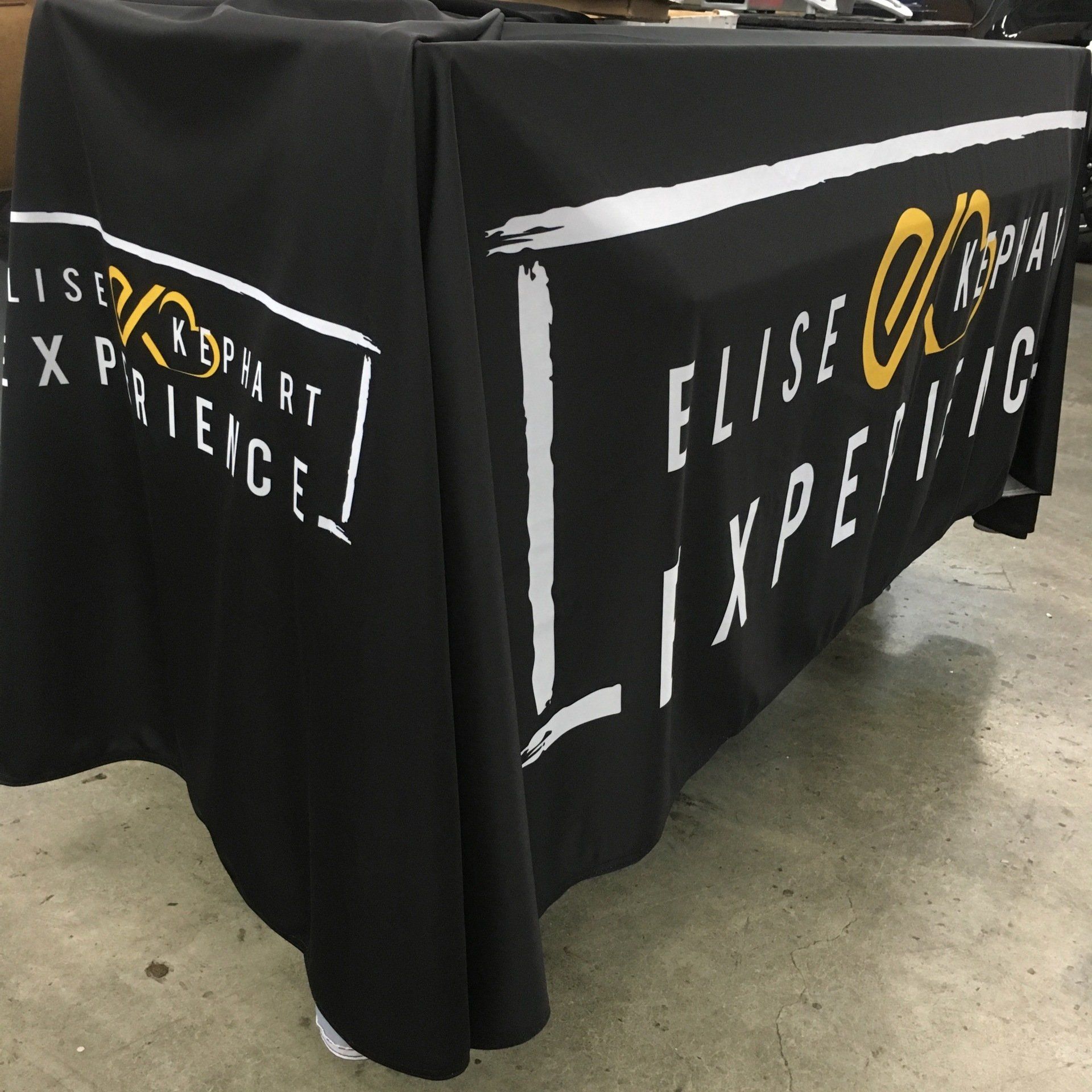 Black tablecloths with the “Elise Experience” logo draped over tables on a concrete floor.