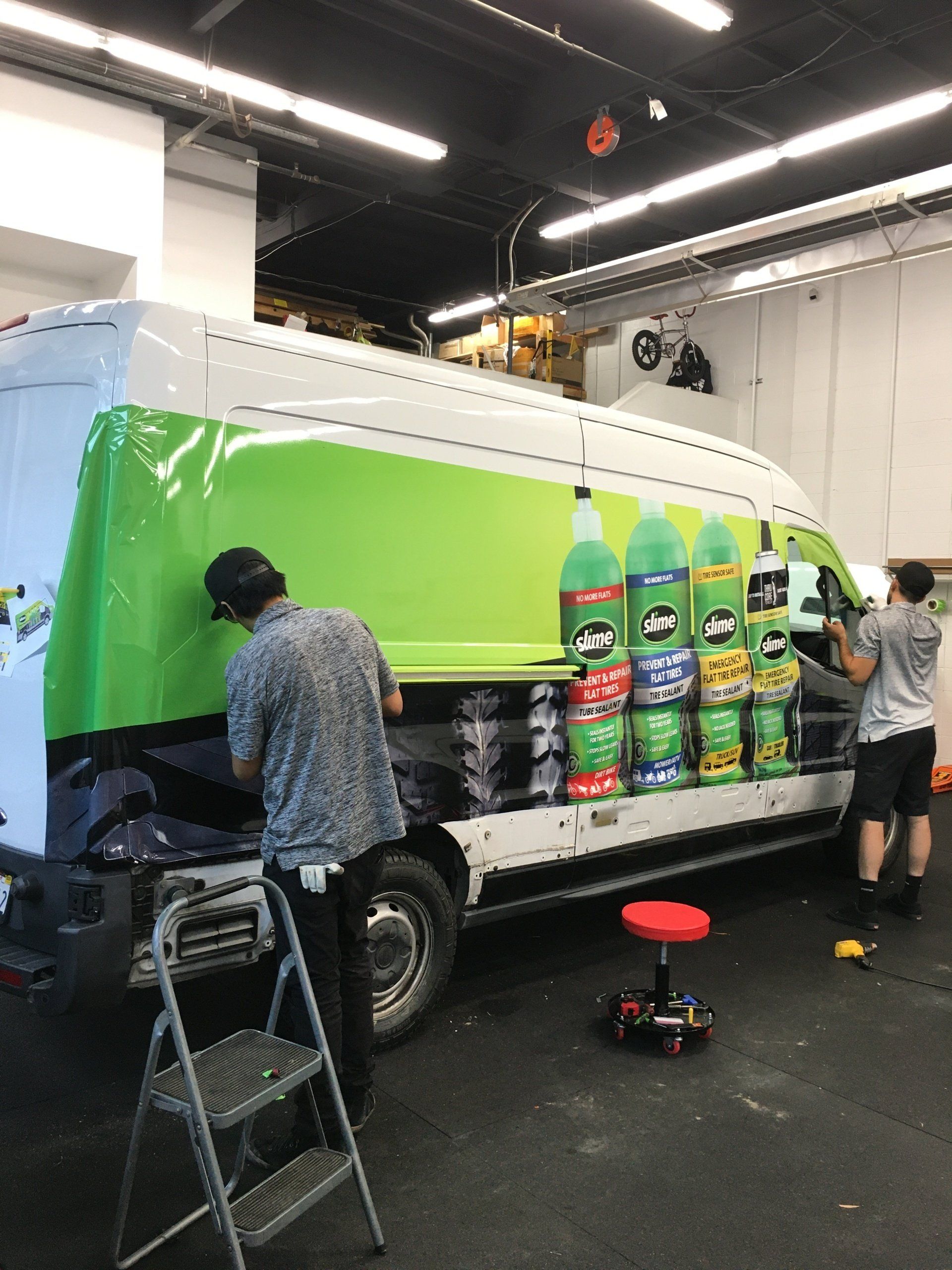Two people wrap a van with a bright green ad inside a workshop, with product graphics on the side.