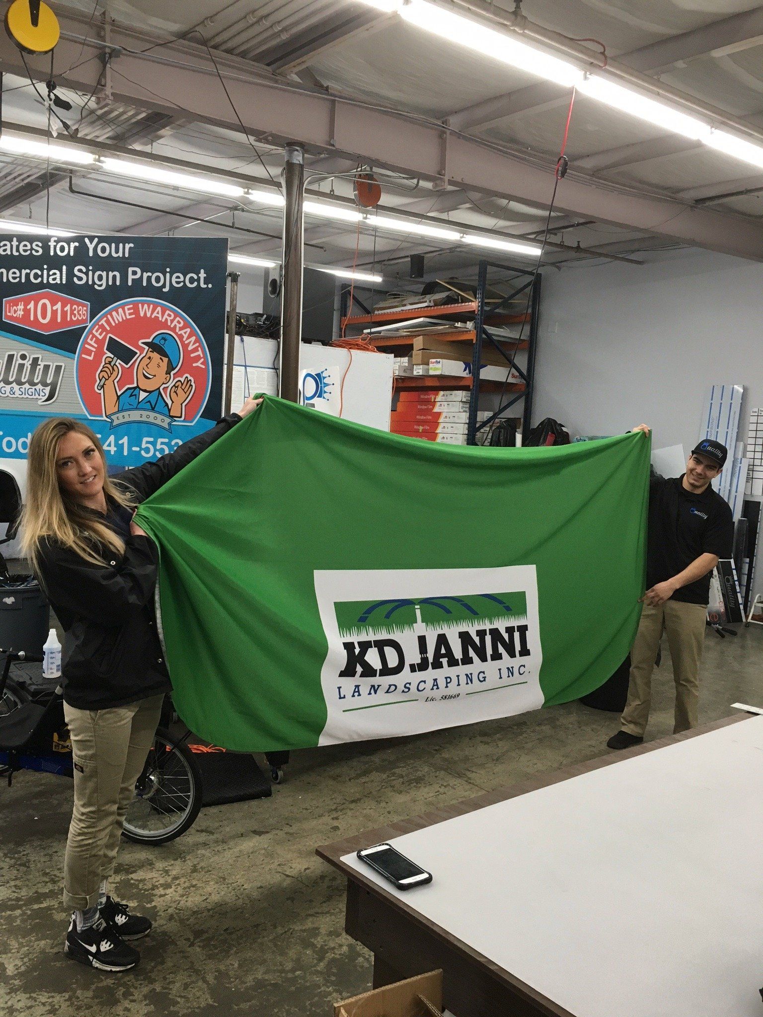 People holding a green cover over a cart in a garage, with a KOJANNI sign visible.