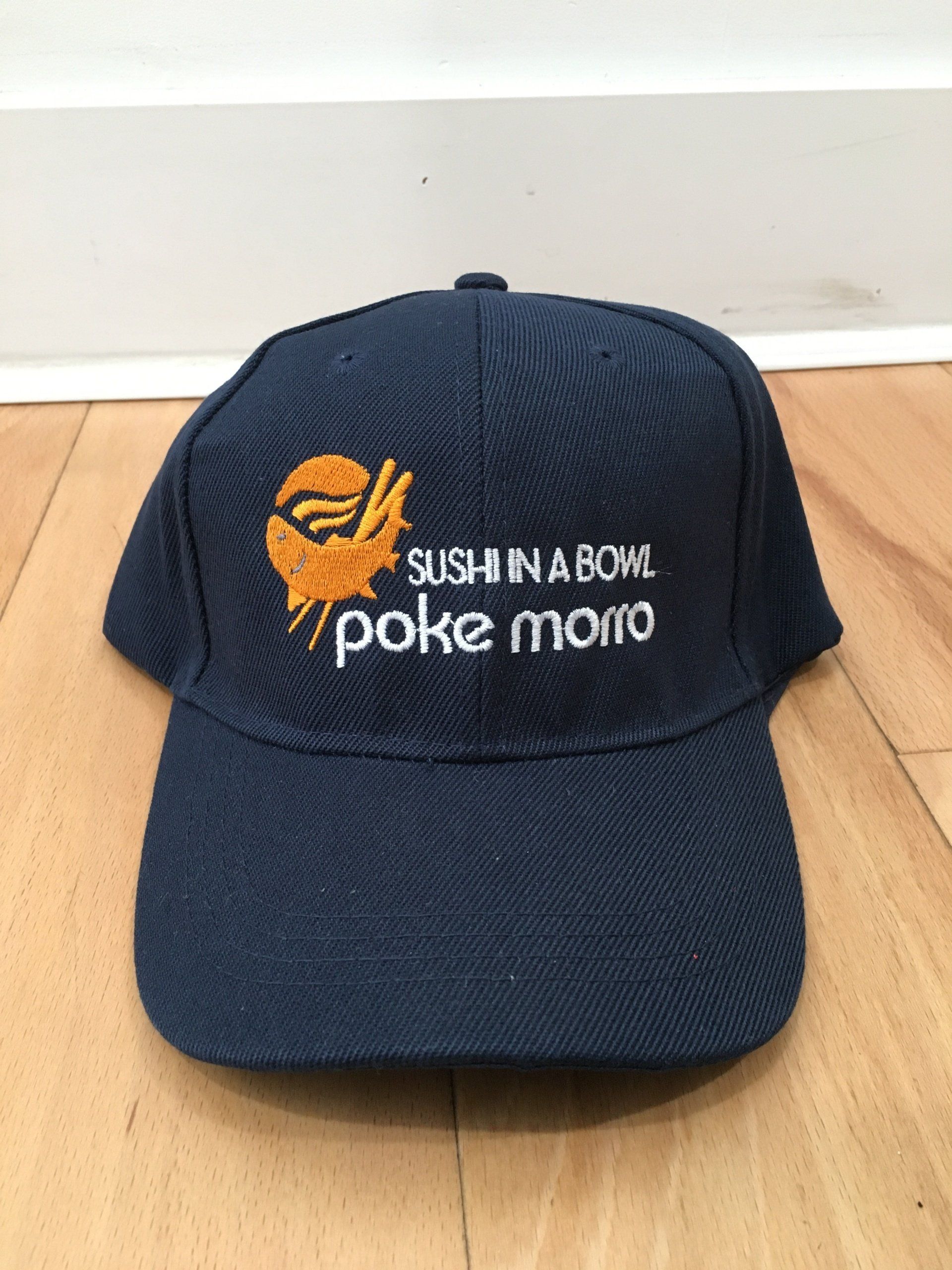 Navy baseball cap with orange graphic and white text reading “SUSHINABOM Poke mon” on a wooden floor.