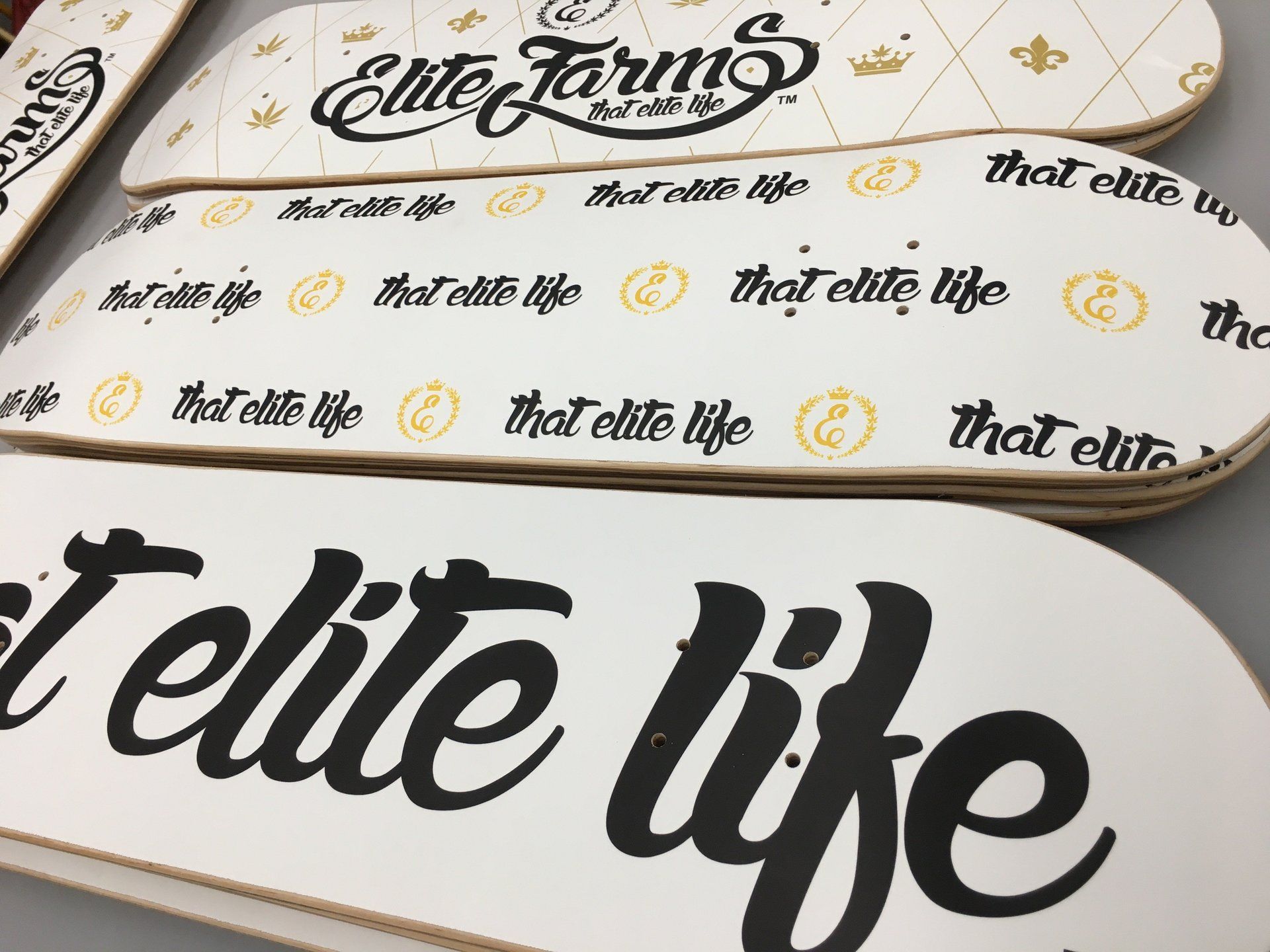 Stacked white skateboard decks with black “elite life” and “Elite Farms” logos on a patterned surface