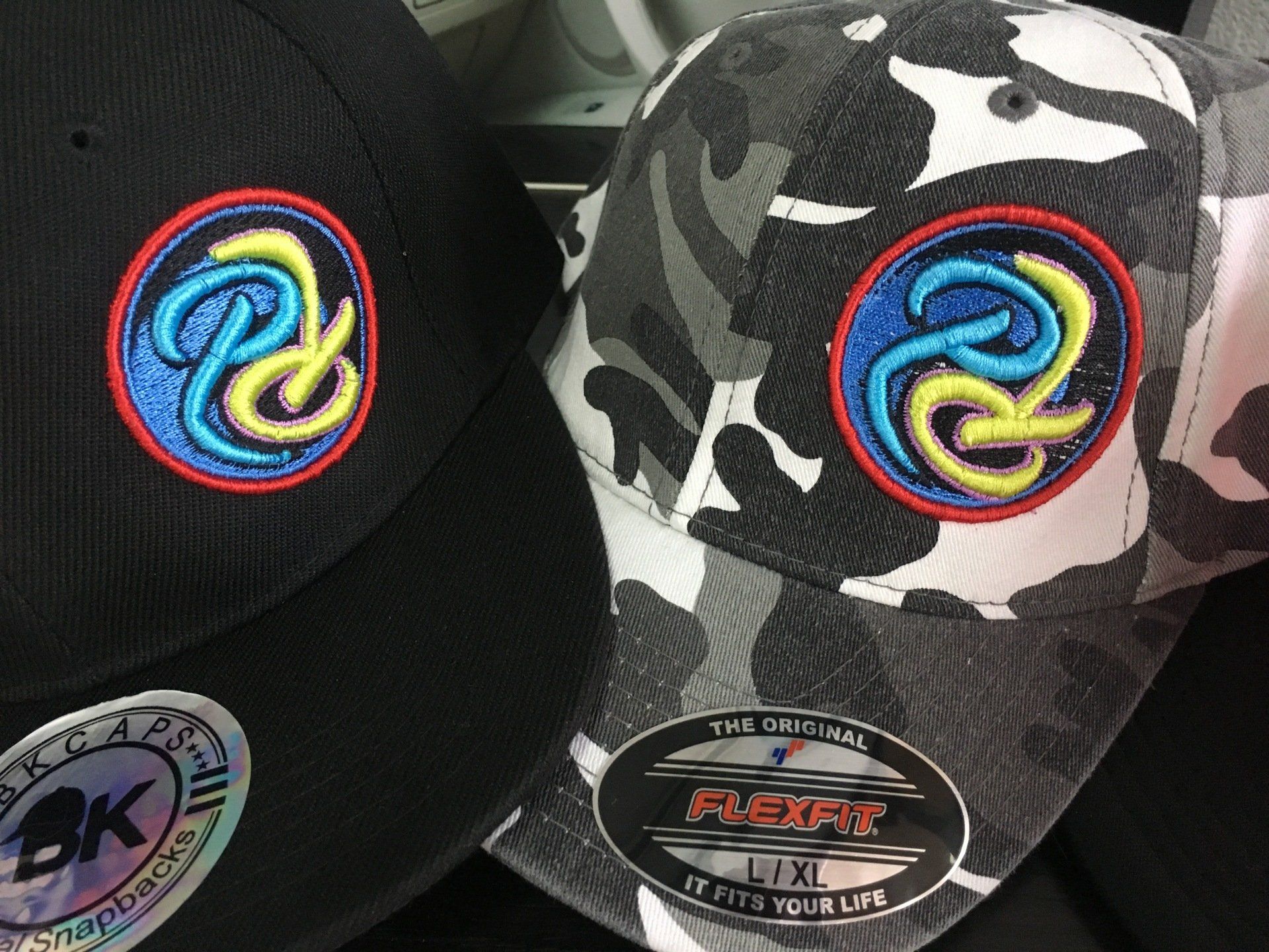 Two branded baseball caps, one black and one camo, each with a colorful embroidered logo.