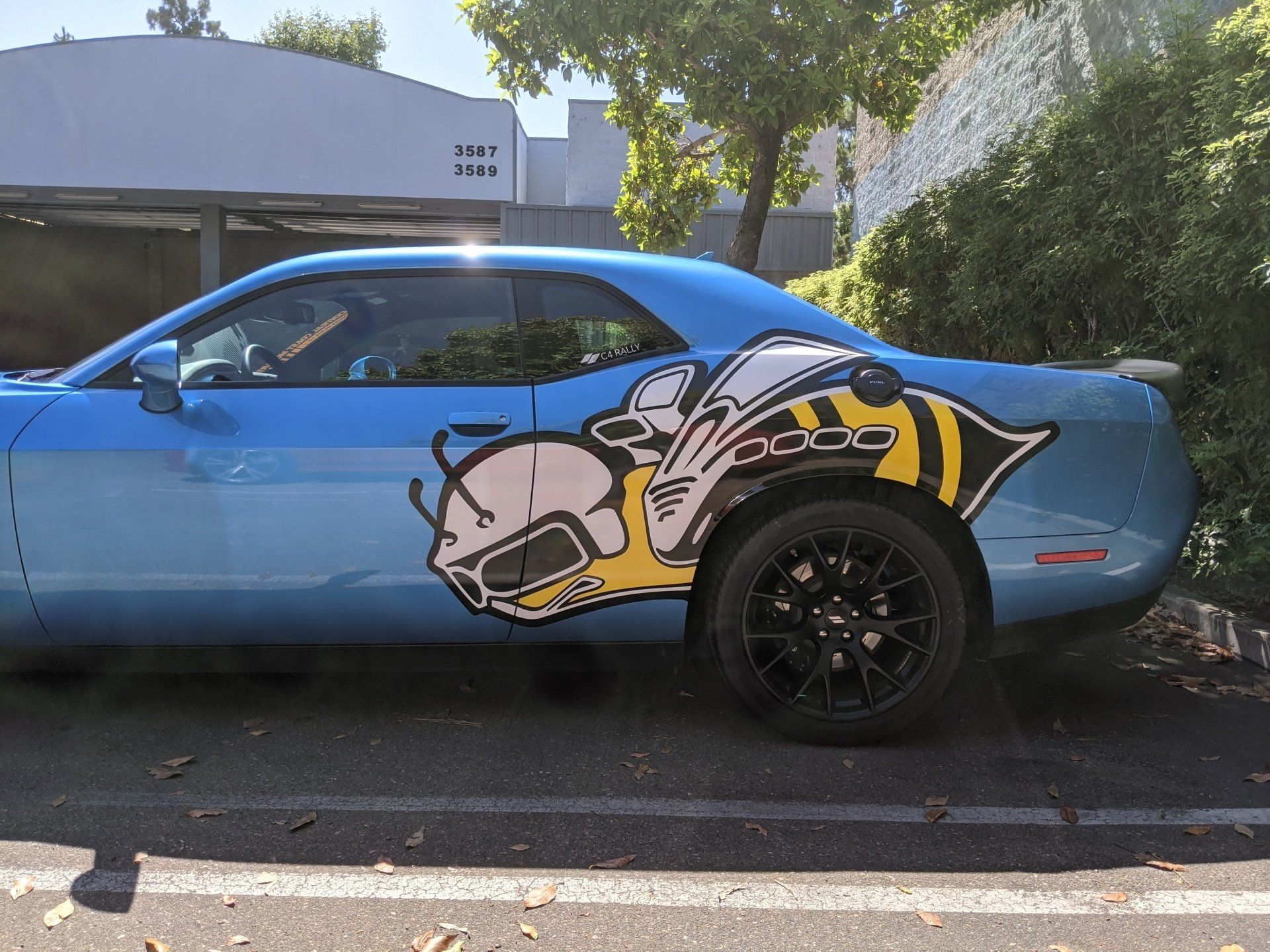 Blue sports car with a large cartoon decal parked on a street beside a wall and trees