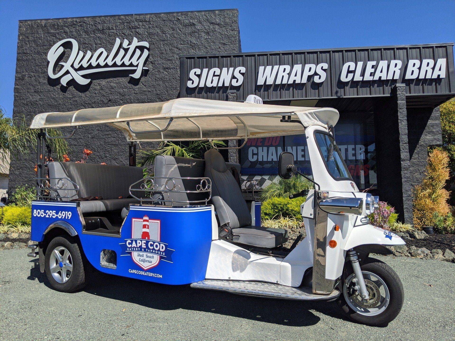 Blue-and-white custom tuk-tuk parked in front of a sign shop with “SIGNS WRAPS CLEAR BRA” signage