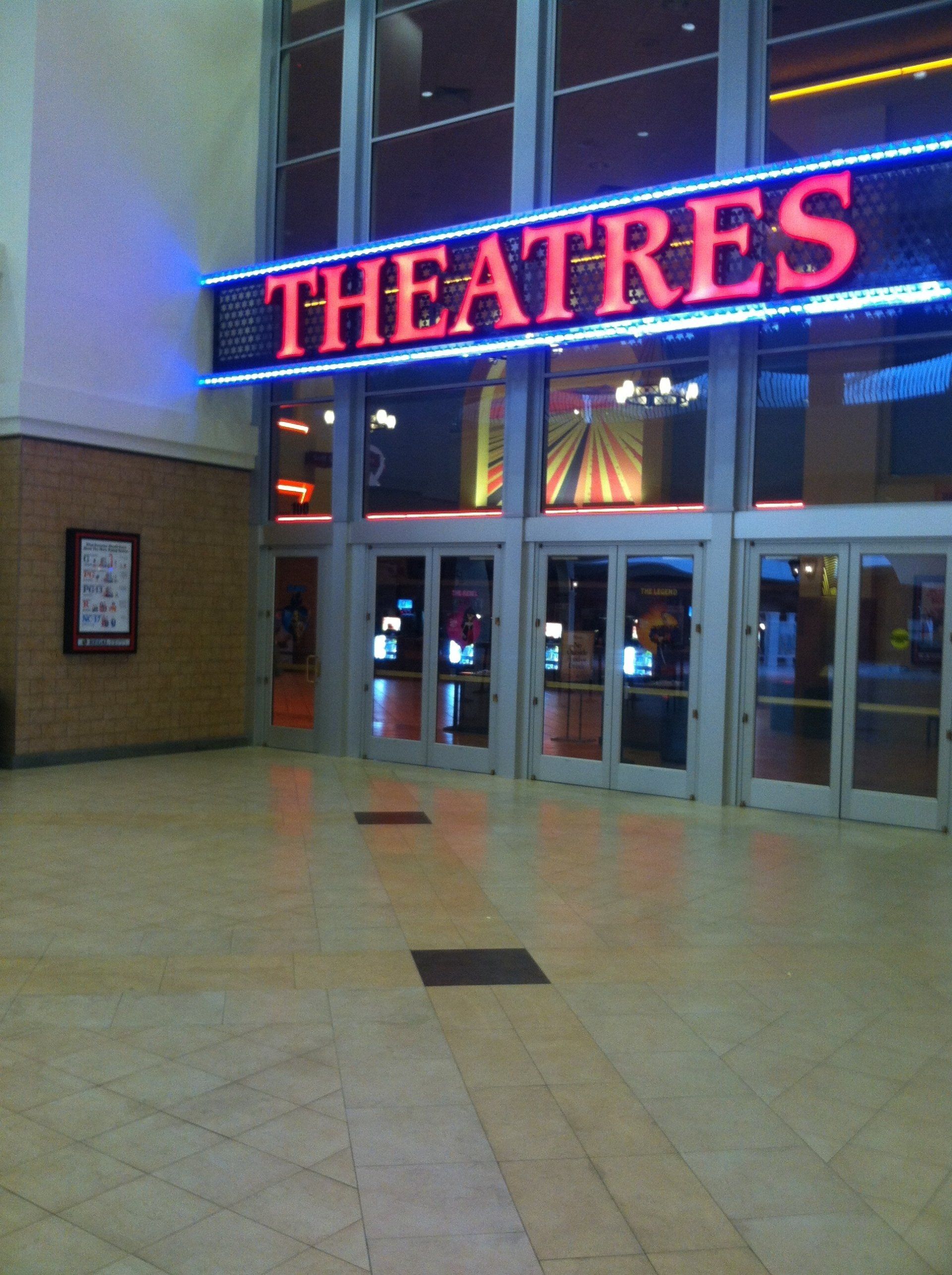 Entrance to a movie theater with a red neon “THEATRES” sign and glass doors in a mall lobby