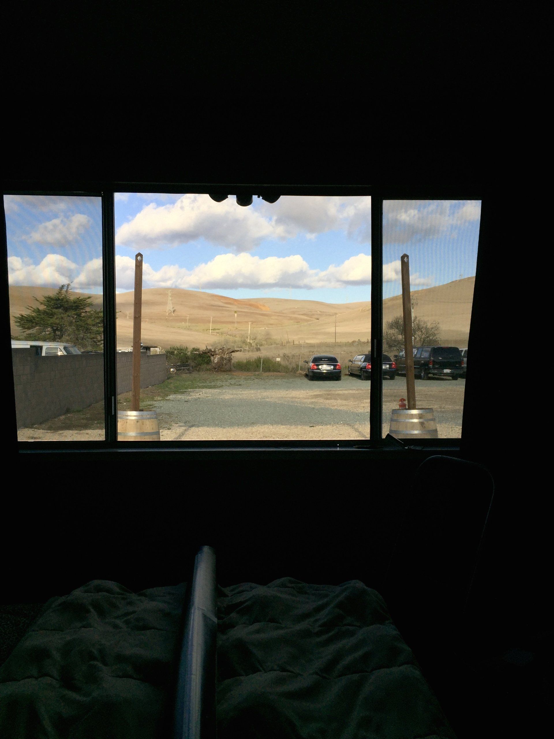 View from a dark room through three windows to a grassy valley, hills, and parked cars under a cloudy blue sky