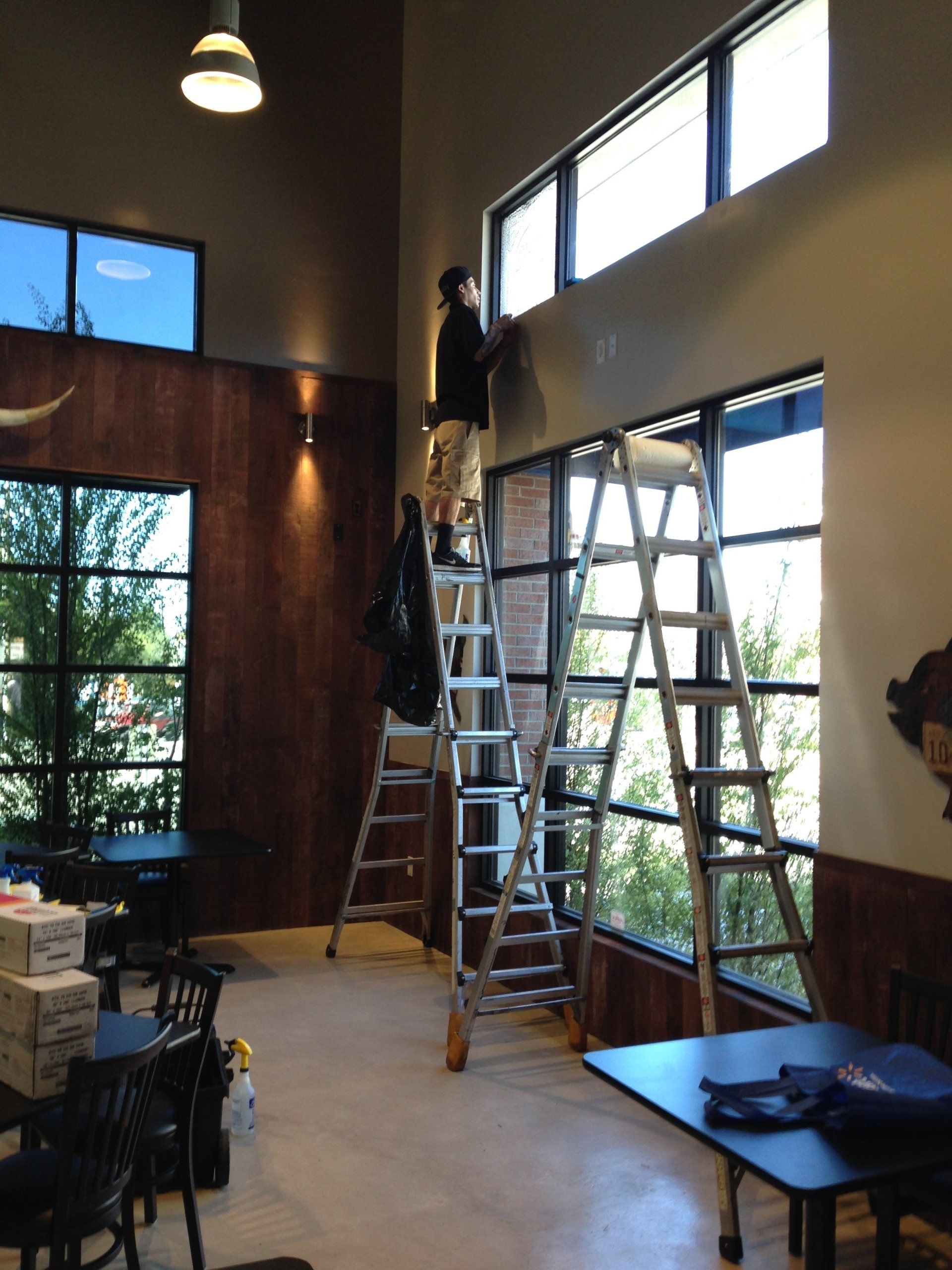 Tall room with ladders set up by large windows and a person working near the ceiling, likely cleaning or painting.