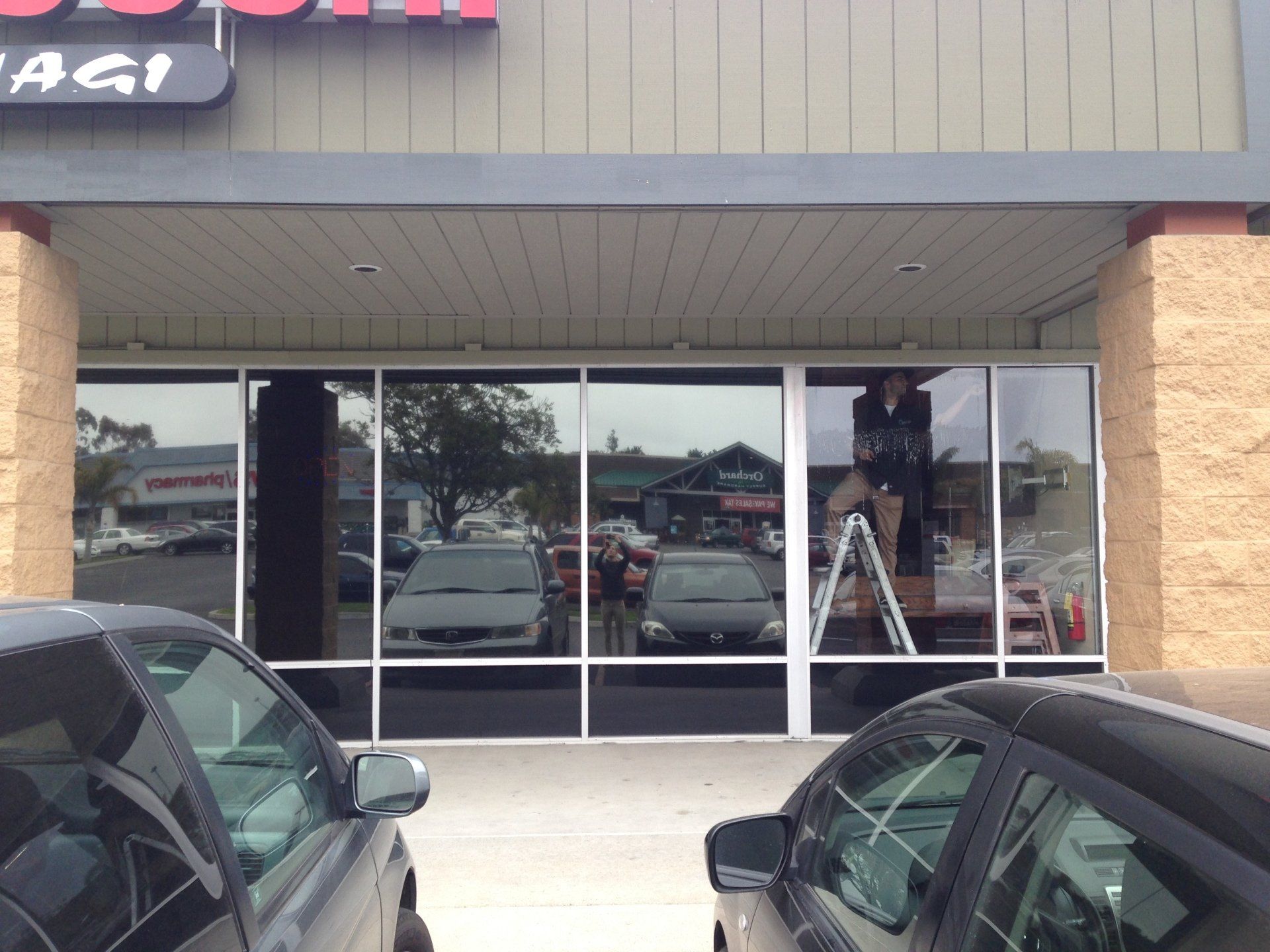 Storefront with dark-tinted glass doors and windows, parked cars in front, and a ladder visible inside.