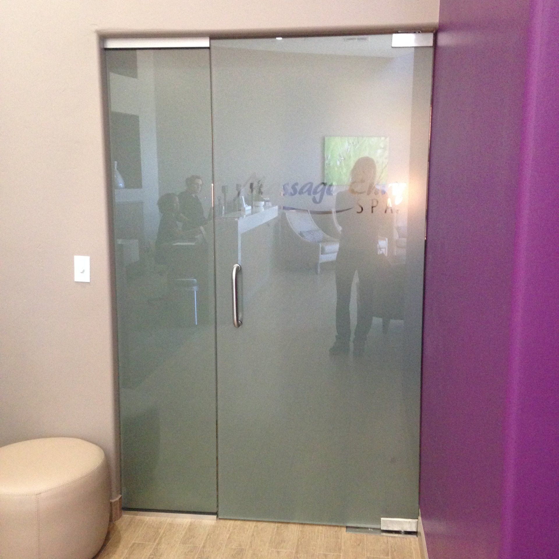 Frosted glass office door with metal handle, reflected room behind, beside a purple wall and beige stool.