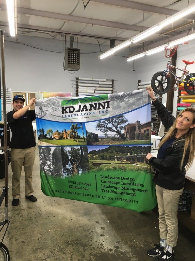 Two people hold a colorful K. D. Janni banner in a workshop garage.