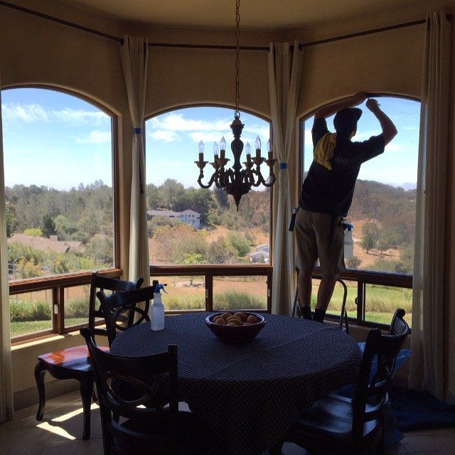 Person hanging curtains in a sunlit dining room with a round table, chandelier, and mountain view.