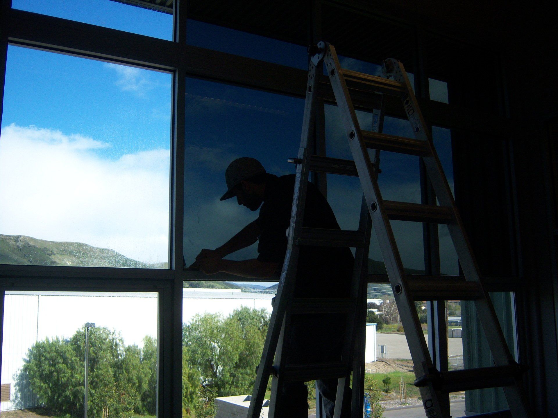Person silhouetted on a ladder working on a large window in a room with a bright blue sky outside.