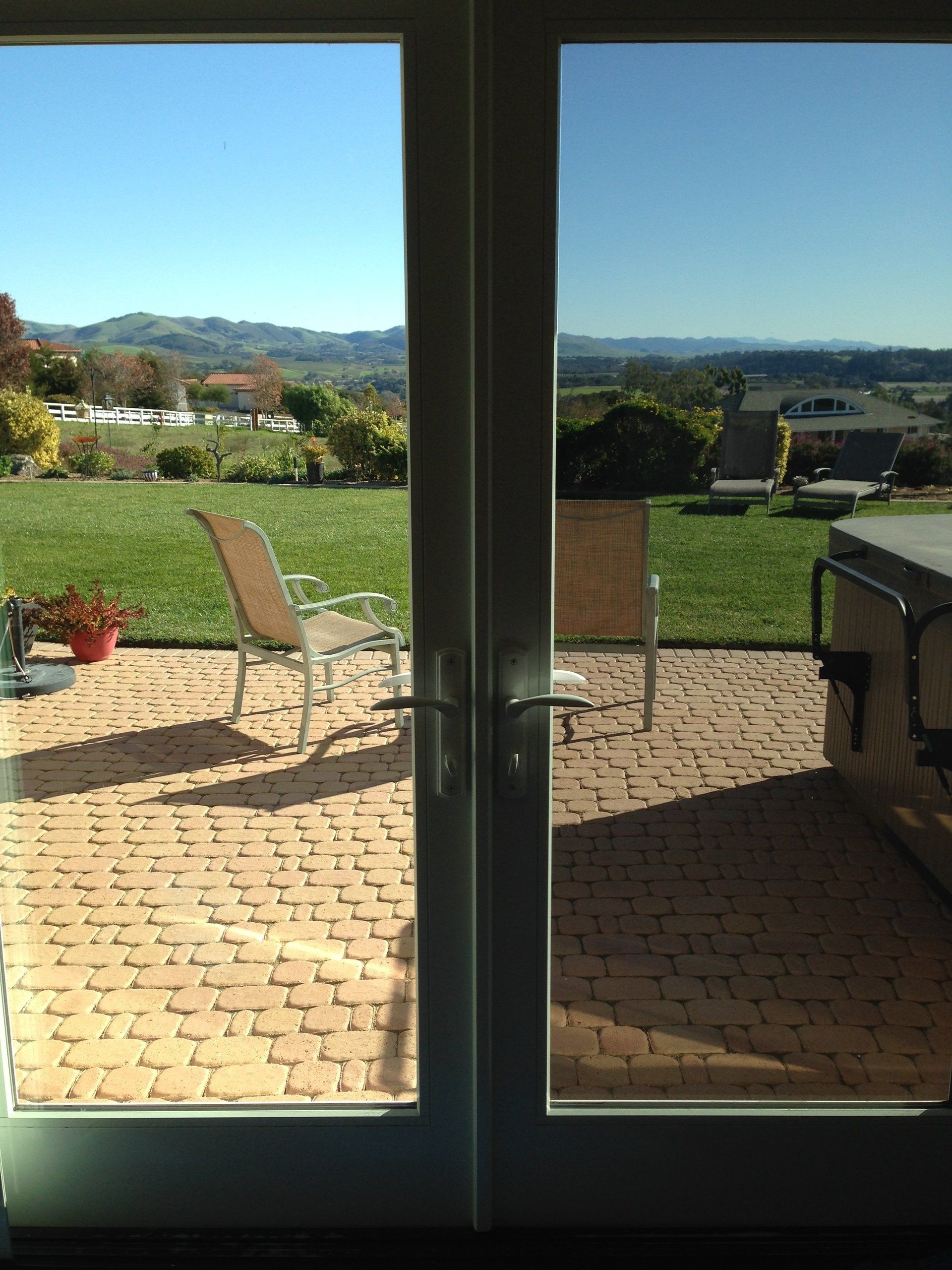 Patio viewed through glass doors, with outdoor chairs, potted plants, and a bright green landscape beyond.
