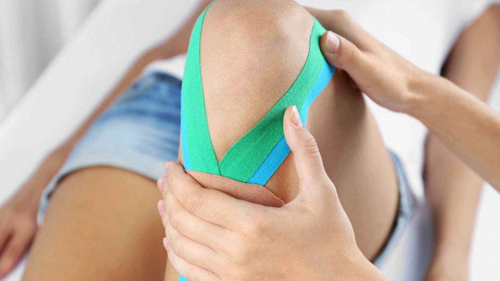 Ginocchio di una persona con nastro kinesiologico verde e blu, toccato dalle mani.